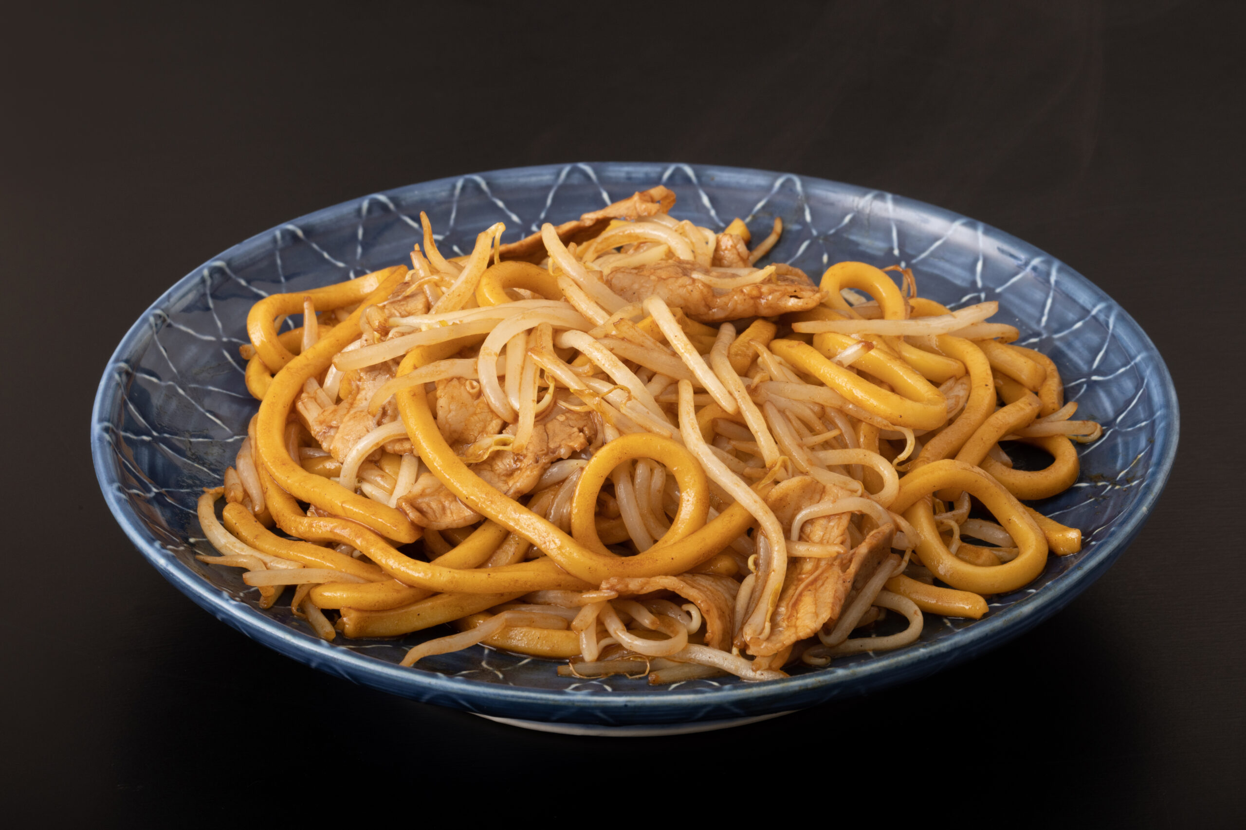Blue ceramic bowl of stir-fried noodles with pork and onions on a dark background, close-up.