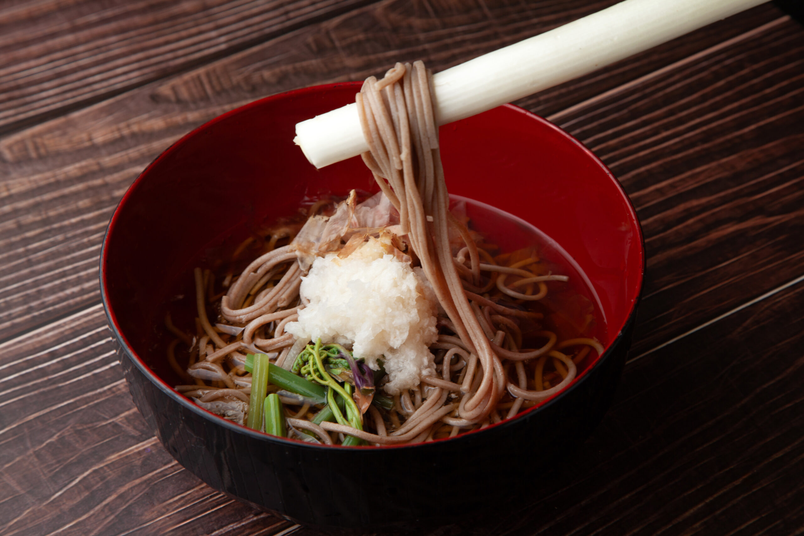 Bowl of soba noodles with grated daikon radish and green onions in a red bowl on a dark wooden table, with chopsticks lifting noodles.