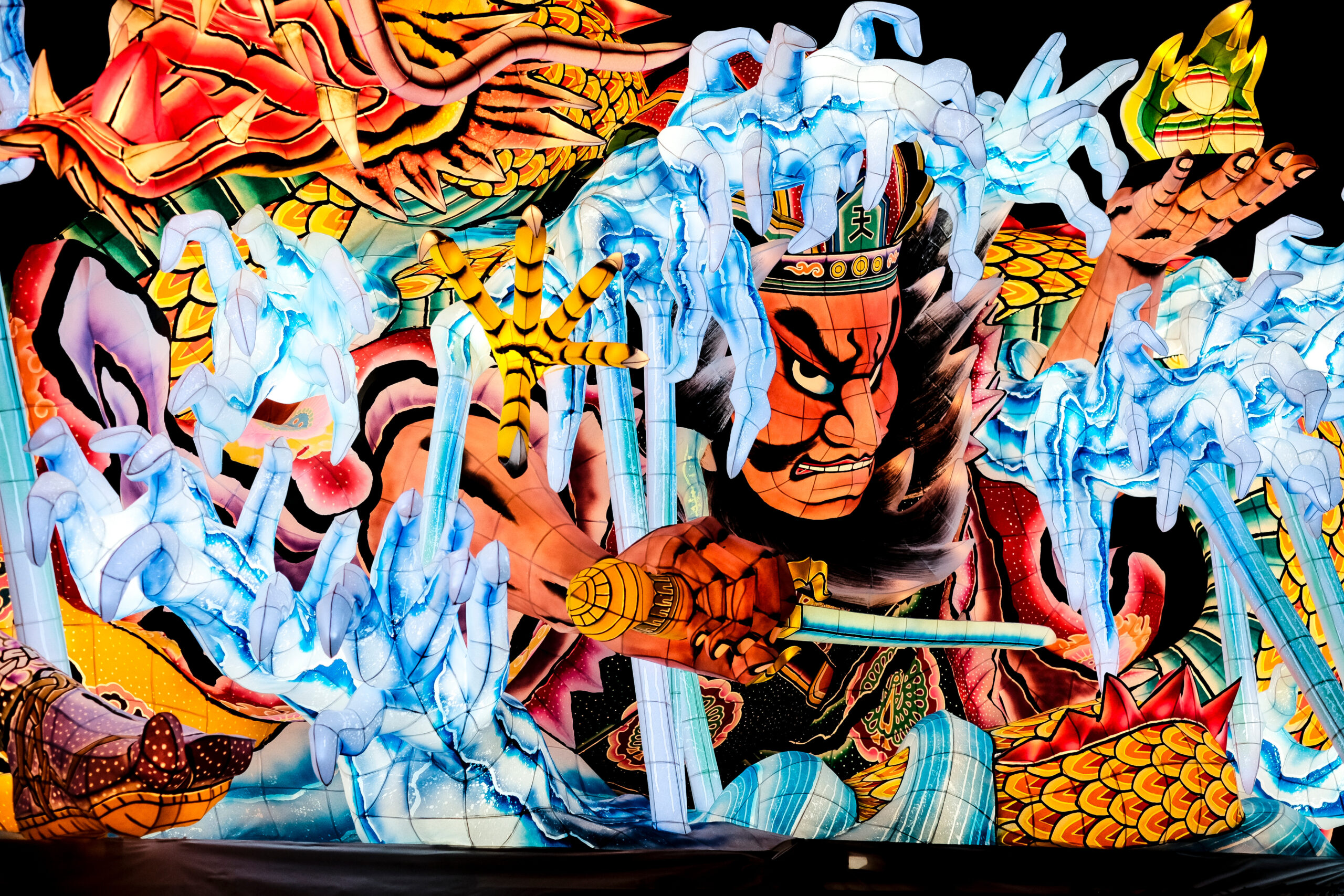 Colorful illuminated dragon lantern sculpture with red, orange, and blue scales and pale blue clawed hands reaching through a dark background at night.