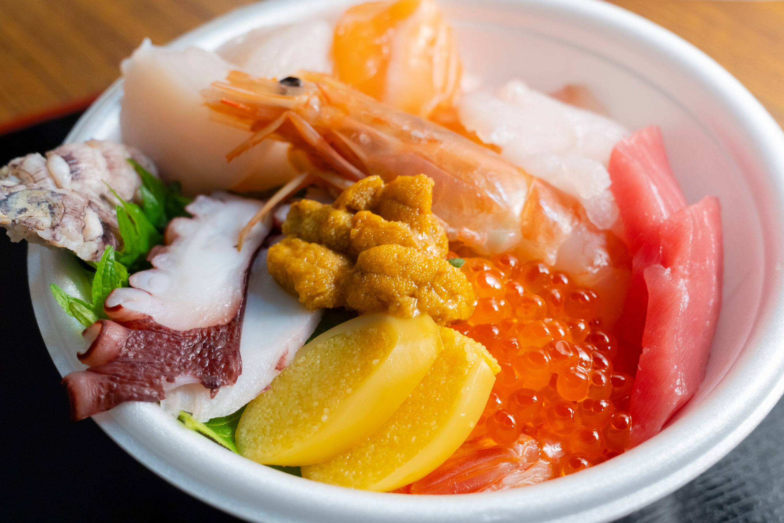 Assorted sashimi bowl with shrimp, tuna, octopus, sea urchin, salmon roe, lemon slice, and greens in a white bowl