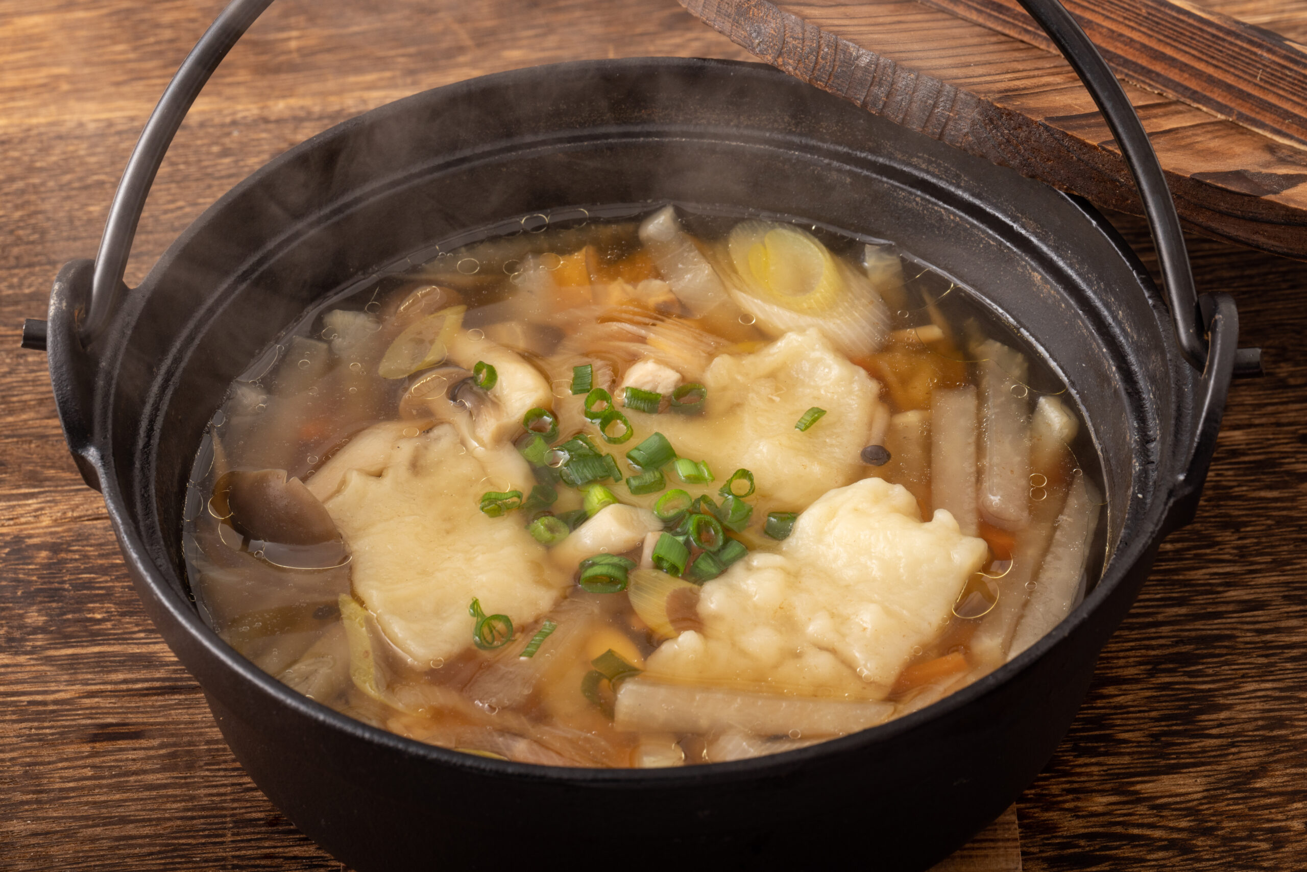 Steaming bowl of dumpling soup with green onions and vegetables in a dark pot on a wooden table