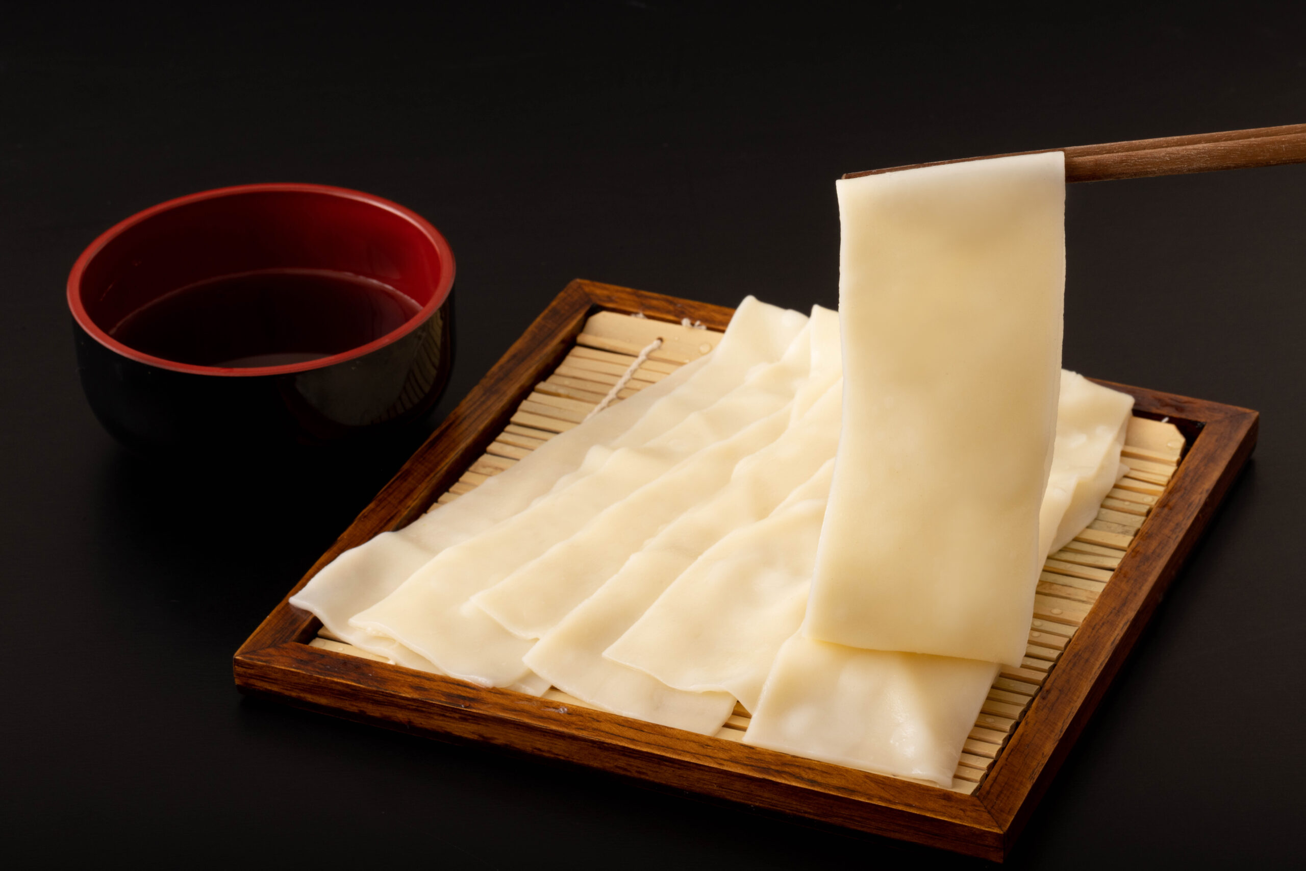 Thin white tofu sheets spread over a wooden bamboo tray with a dark sauce bowl nearby against a black background.