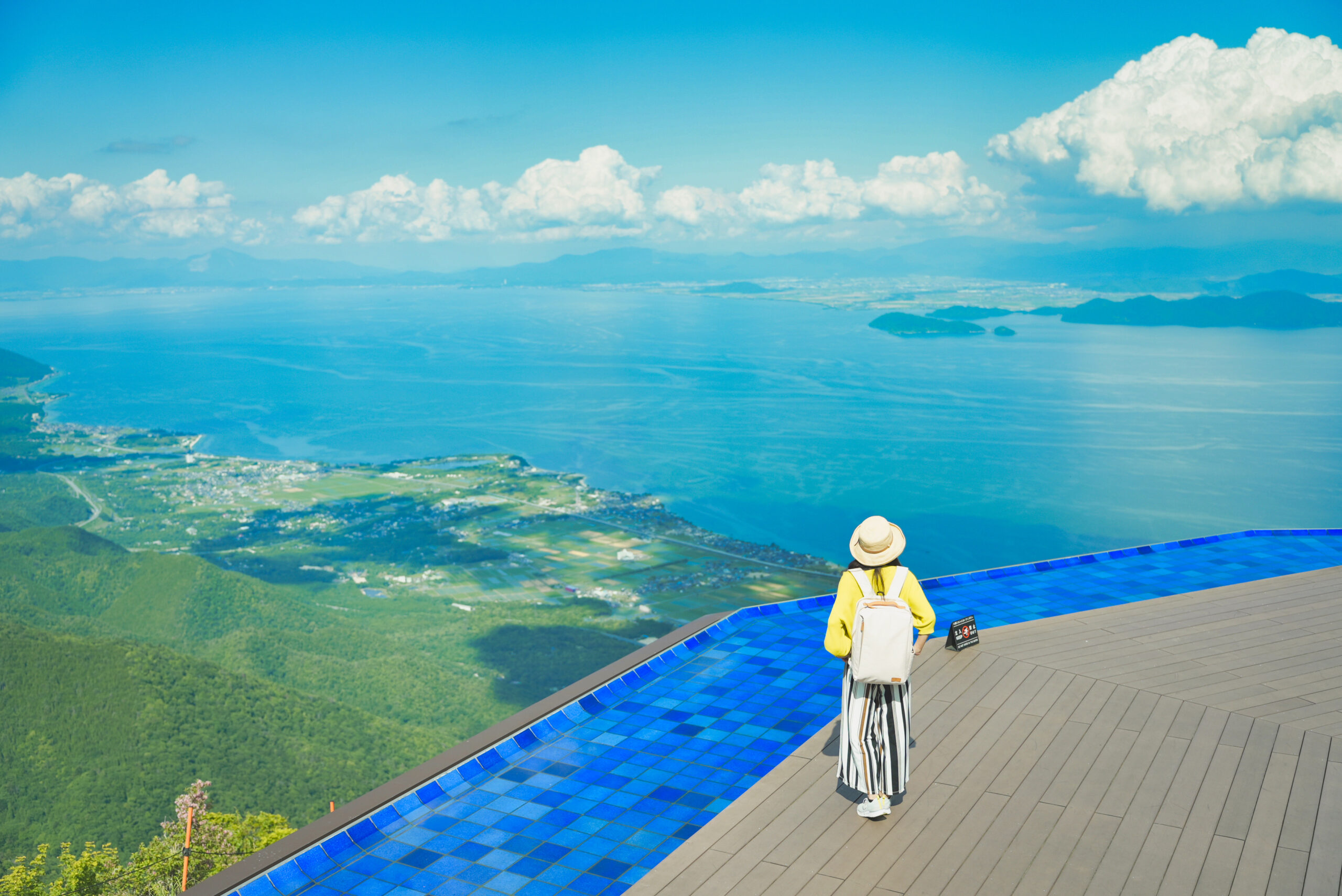 Person wearing a yellow jacket and backpack stands at a blue-tiled infinity pool edge, overlooking a coastal landscape with sea and distant islands.