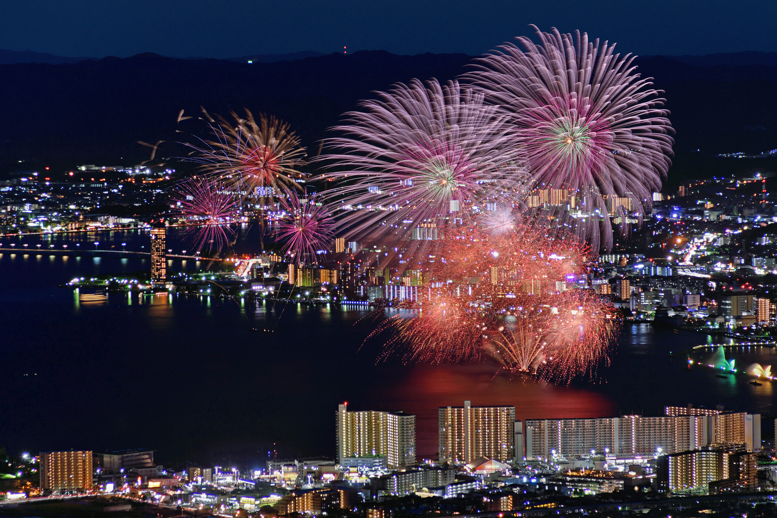 Nighttime city skyline illuminated by colorful fireworks over the harbor, with reflections on the water and illuminated buildings below.