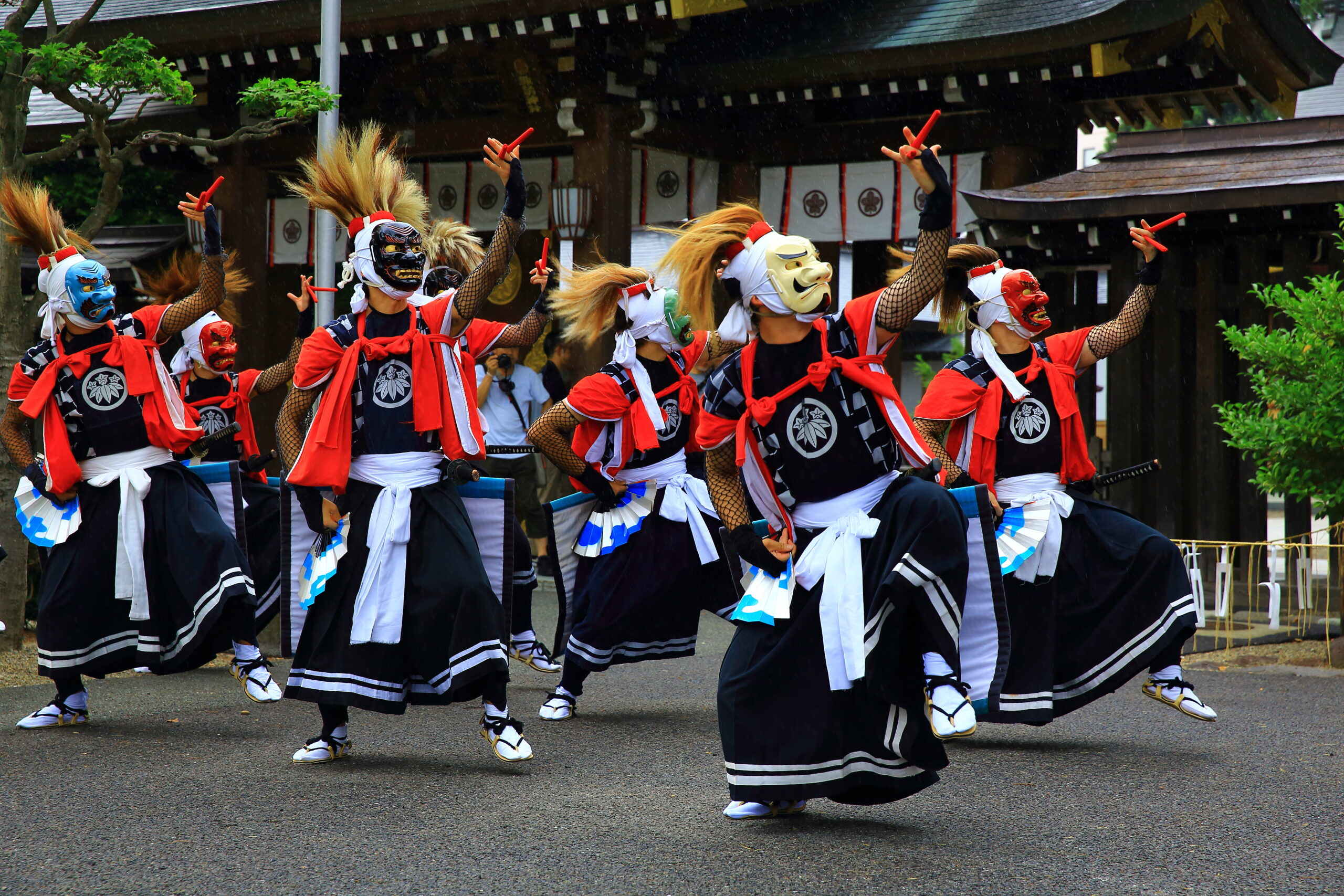 Group of dancers in red, blue, and white costumes with colorful masks performing a festival dance, raising red sticks in front of a shrine.