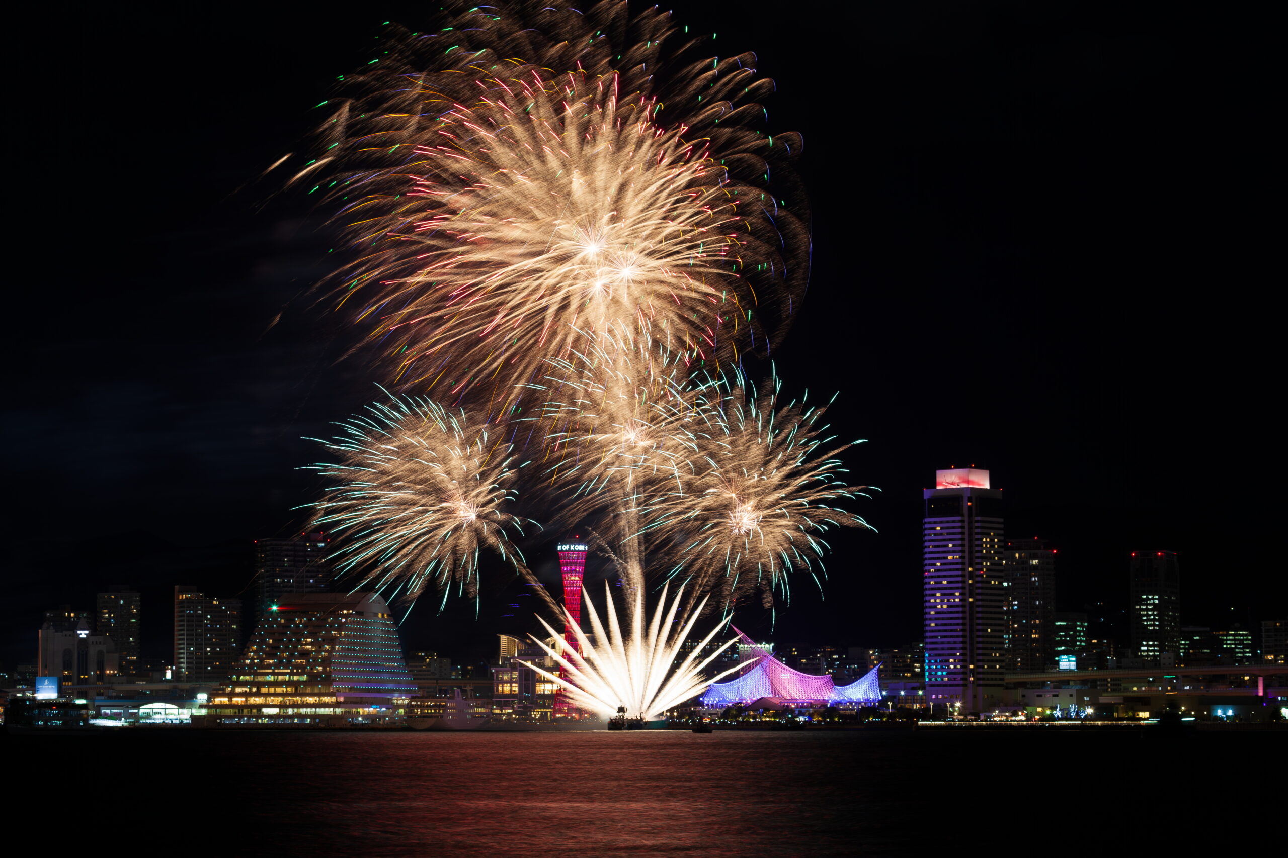 Fireworks explode over a city skyline at night, reflecting on the water below with colorful bursts lighting the buildings.