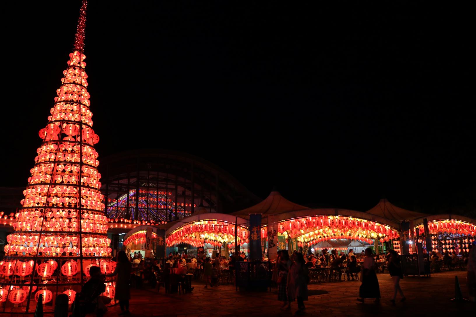 Tall cone-shaped tower of red lanterns at a night festival, with glowing stalls and crowds in the background.