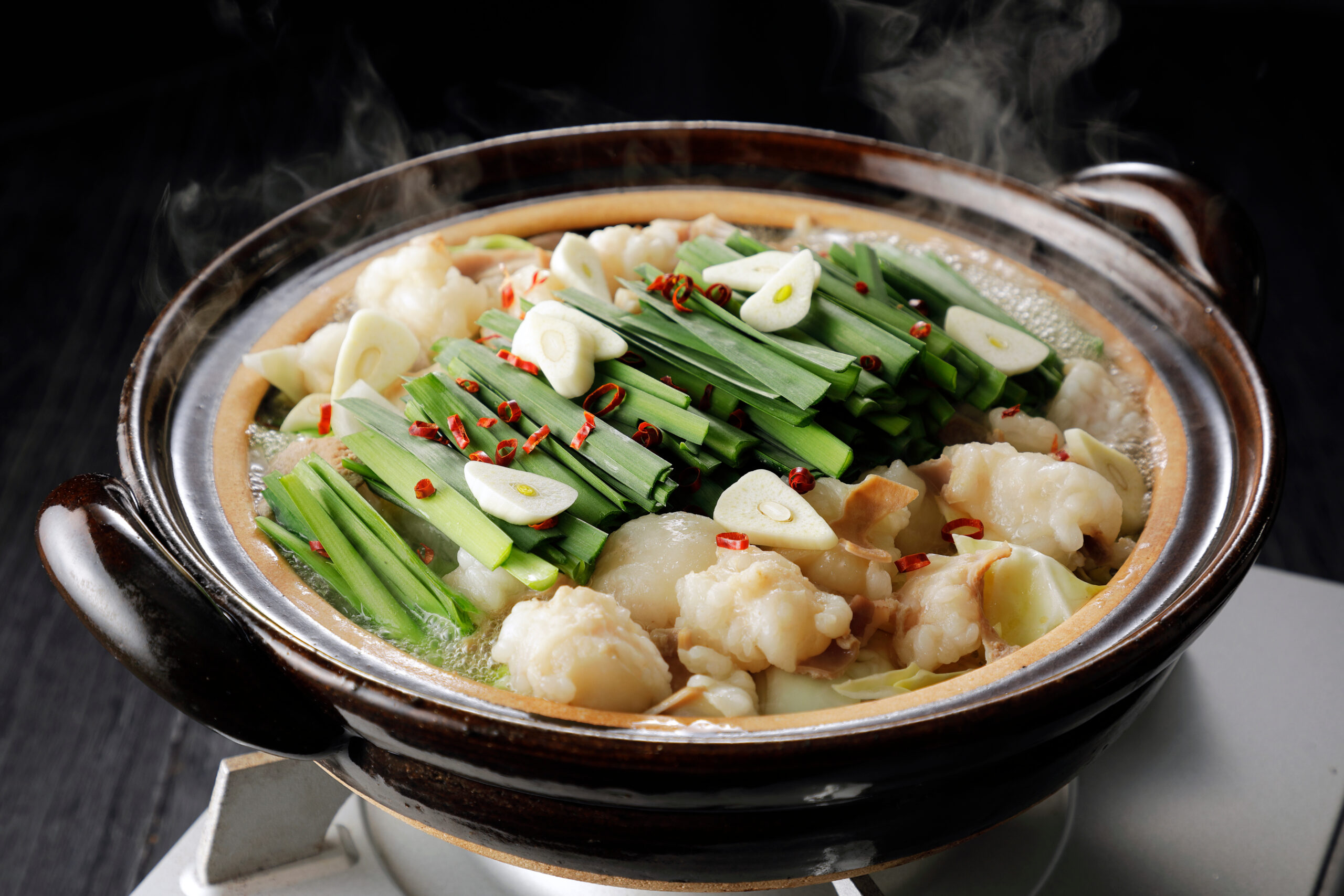 Steaming clay pot with dumplings, scallions, garlic slices, and red chilies in broth.