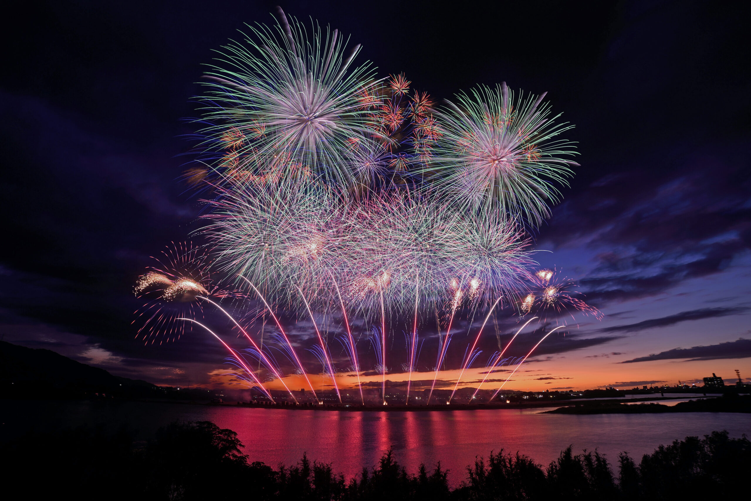 Night sky over a calm river as colorful fireworks explode in multiple bursts above the horizon at dusk.