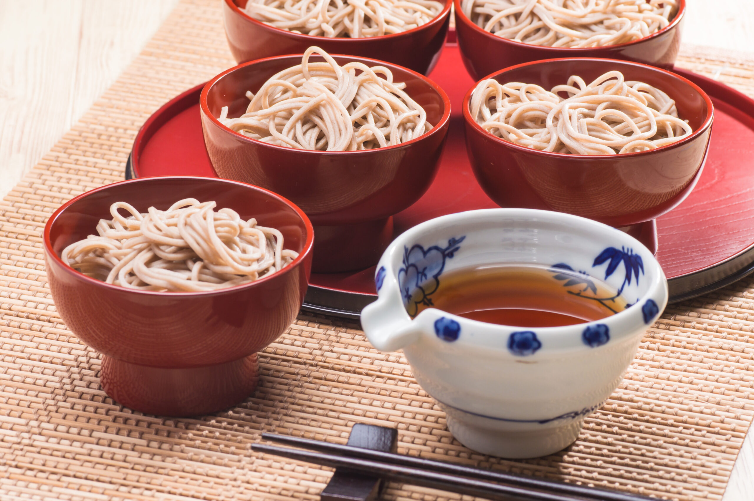Bowl of soba noodles in red lacquer bowls arranged on a bamboo mat with soy sauce cup nearby