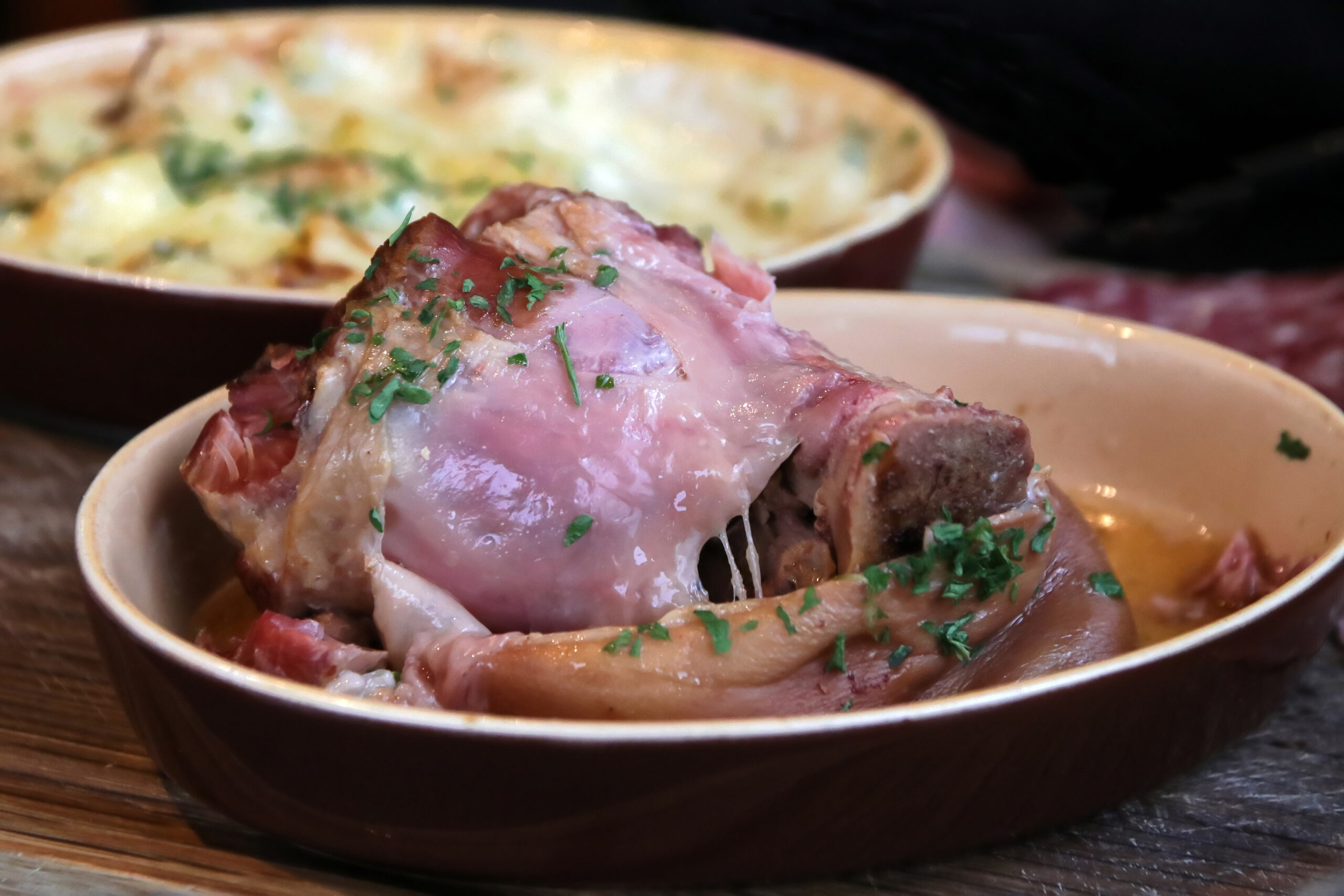 Roasted meat topped with chopped parsley in a ceramic bowl, with a cheesy dish blurred in the background.