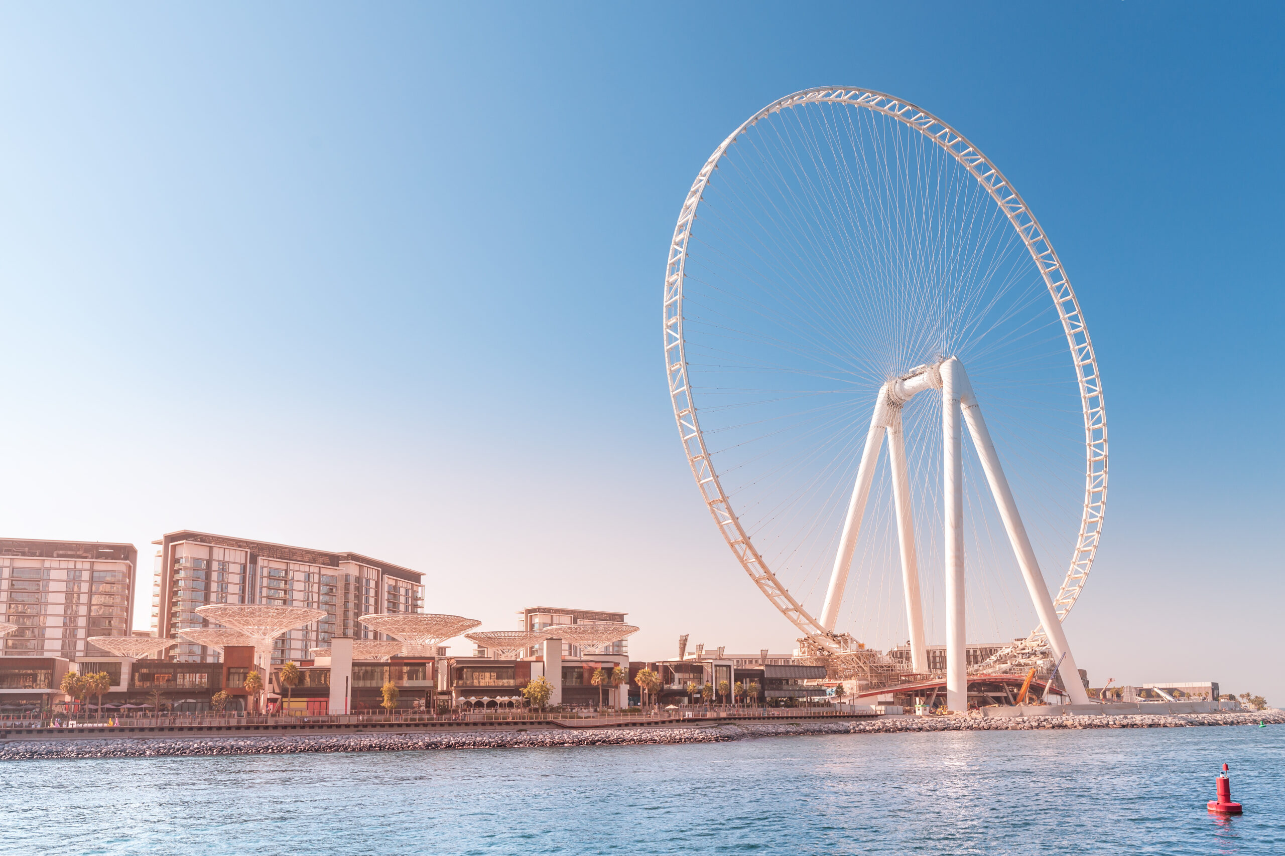Large white Ferris wheel by a waterfront with modern buildings along the shore under a clear blue sky