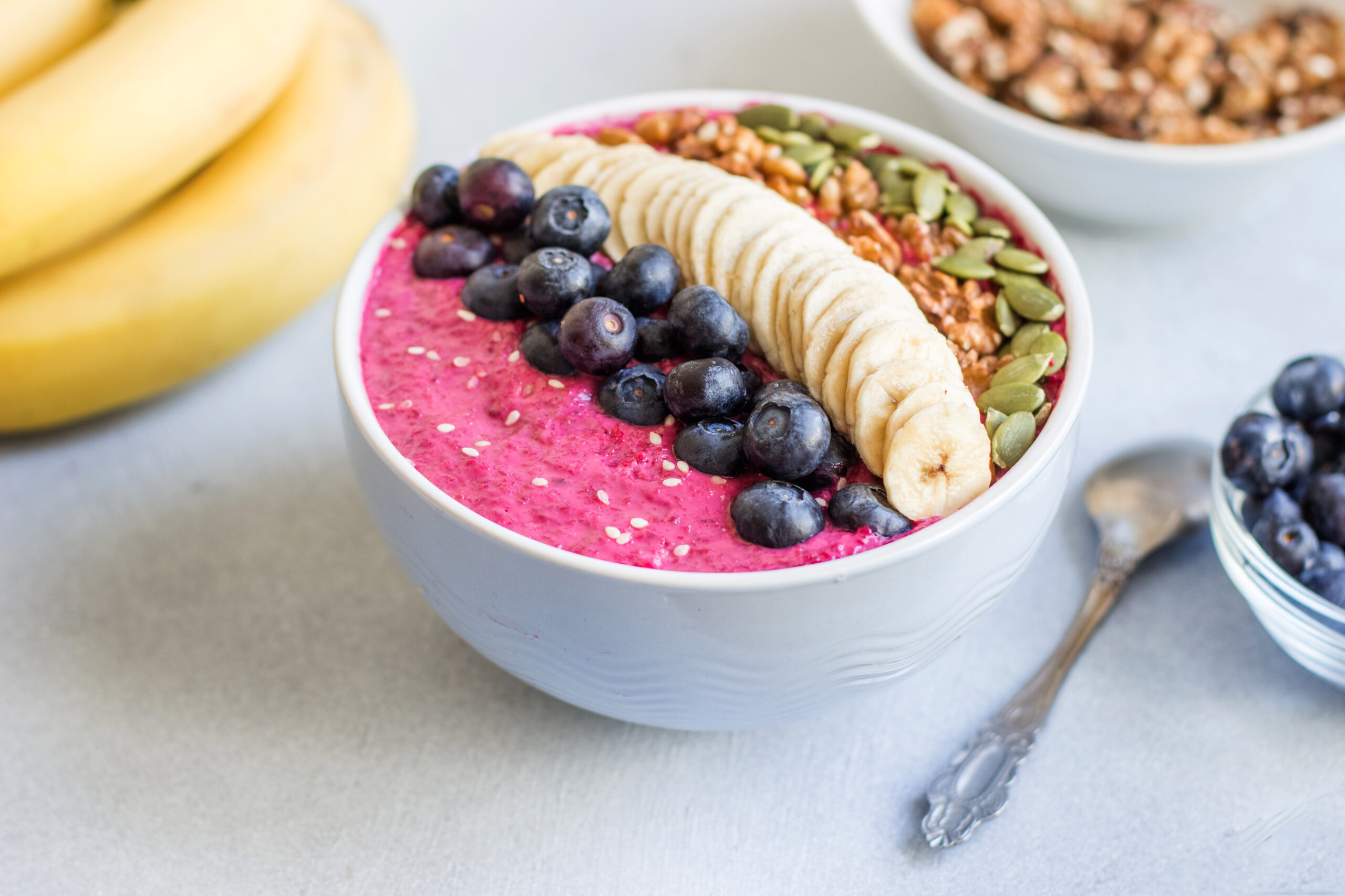 Smoothie bowl with pink base topped with blueberries, banana slices, and seeds on a white bowl.