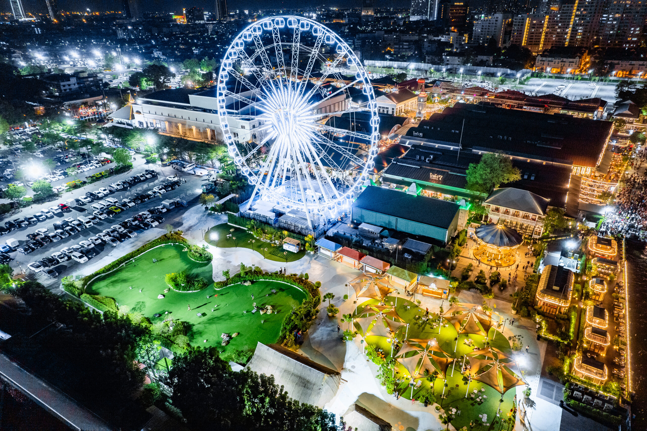 Aerial night view of a brightly lit Ferris wheel at a busy amusement park with surrounding buildings and parking lots.