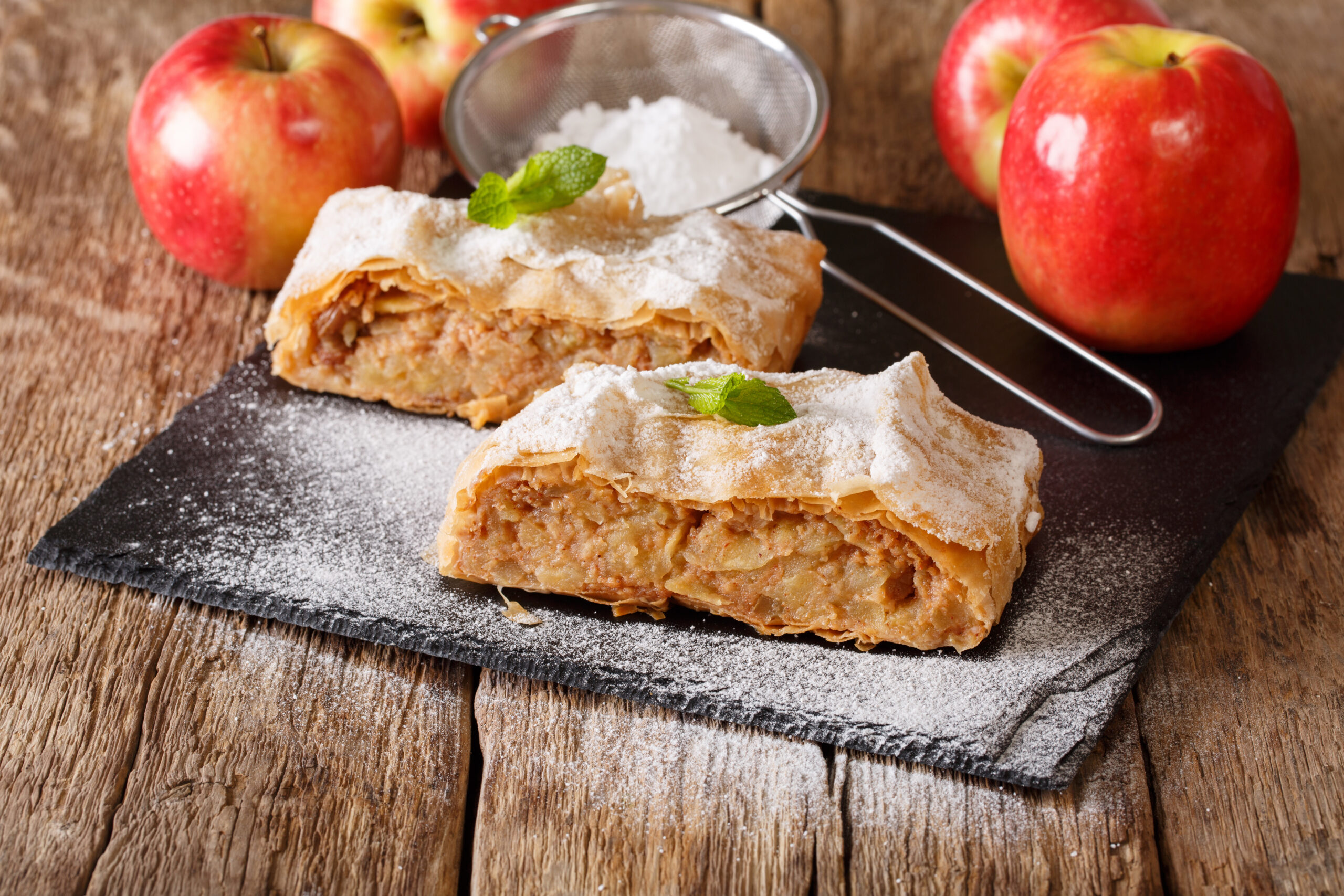 Apple strudel slices on a dark slate board, lightly dusted with powdered sugar and garnished with mint, on a rustic wooden table.
