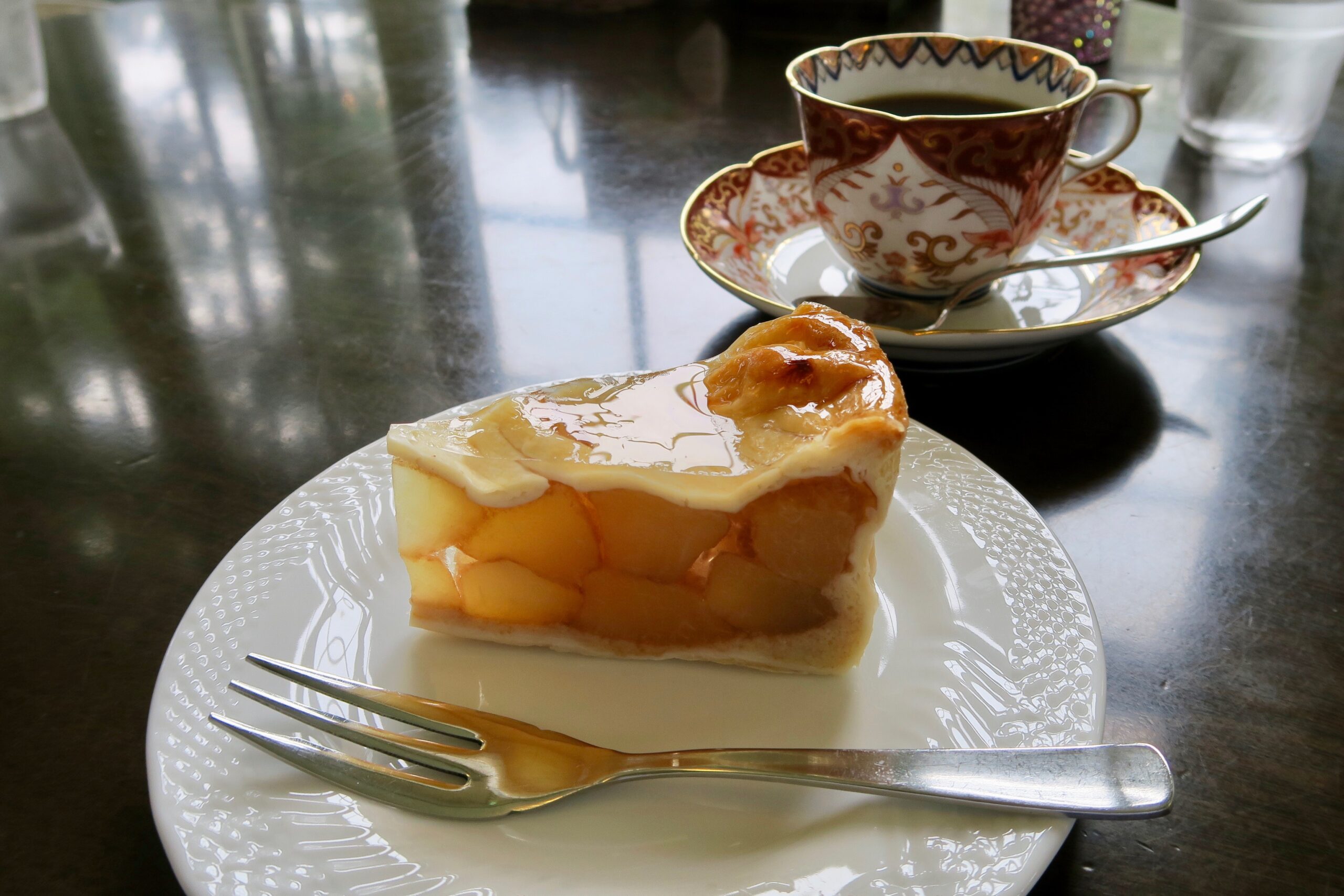 Slice of glazed cake on a white plate with a ornate teacup and saucer in the background on a reflective table surface.