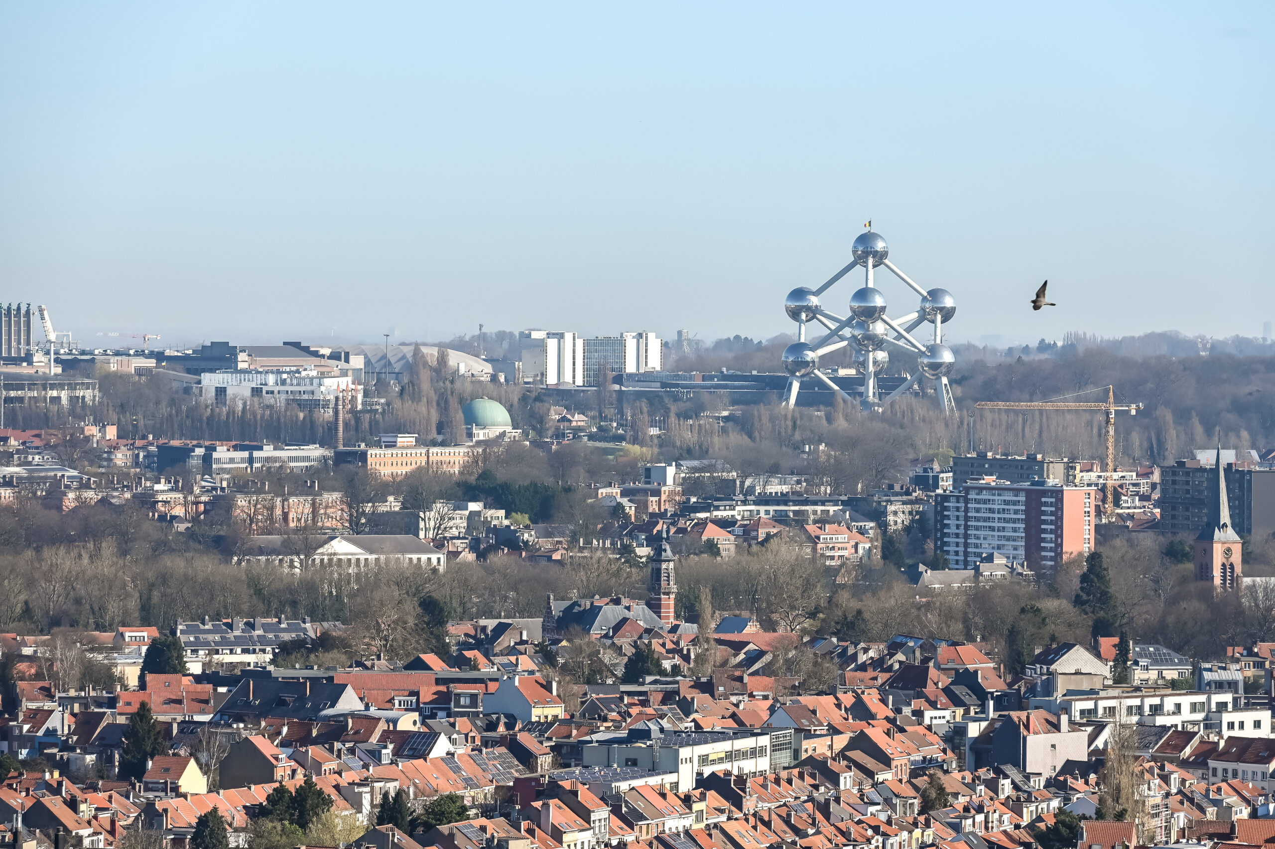 Wide cityscape with red-tiled roofs and the Atomium monument visible in the distance, under a clear blue sky.