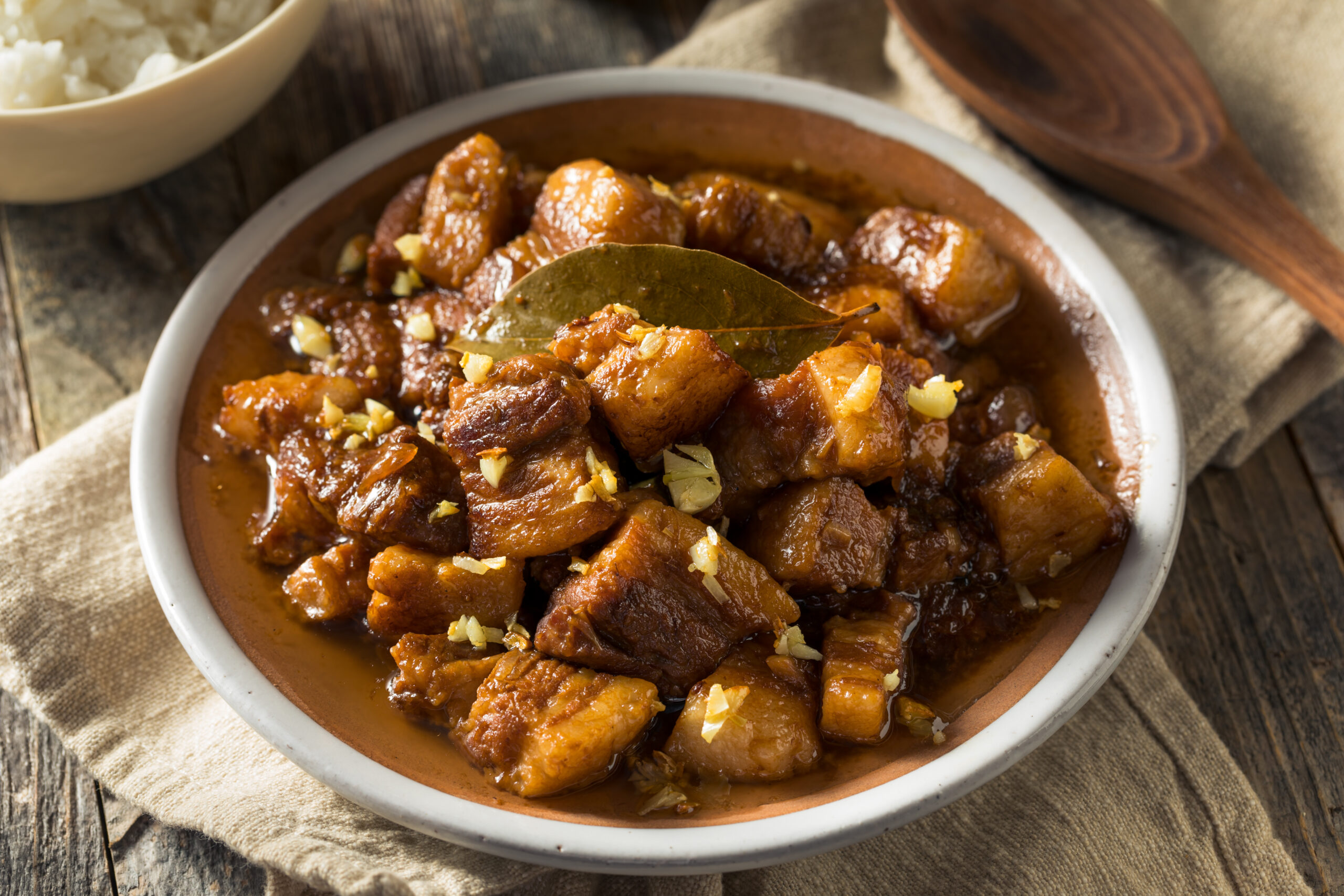 Braised pork chunks in a thick brown sauce with garlic pieces and a bay leaf, served in a white bowl with a side of rice in the background