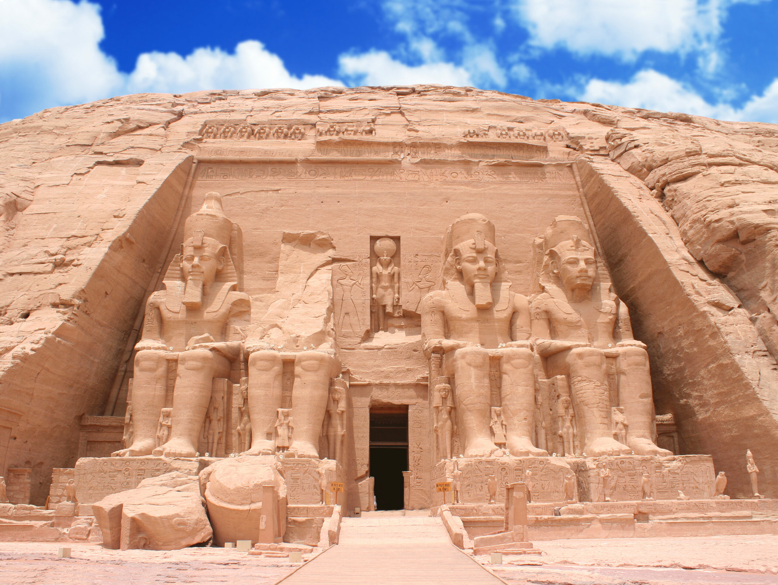 Sand-colored rock-cut temple facade with four colossal Ramesses II statues at the entrance under a blue sky, Abu Simbel.