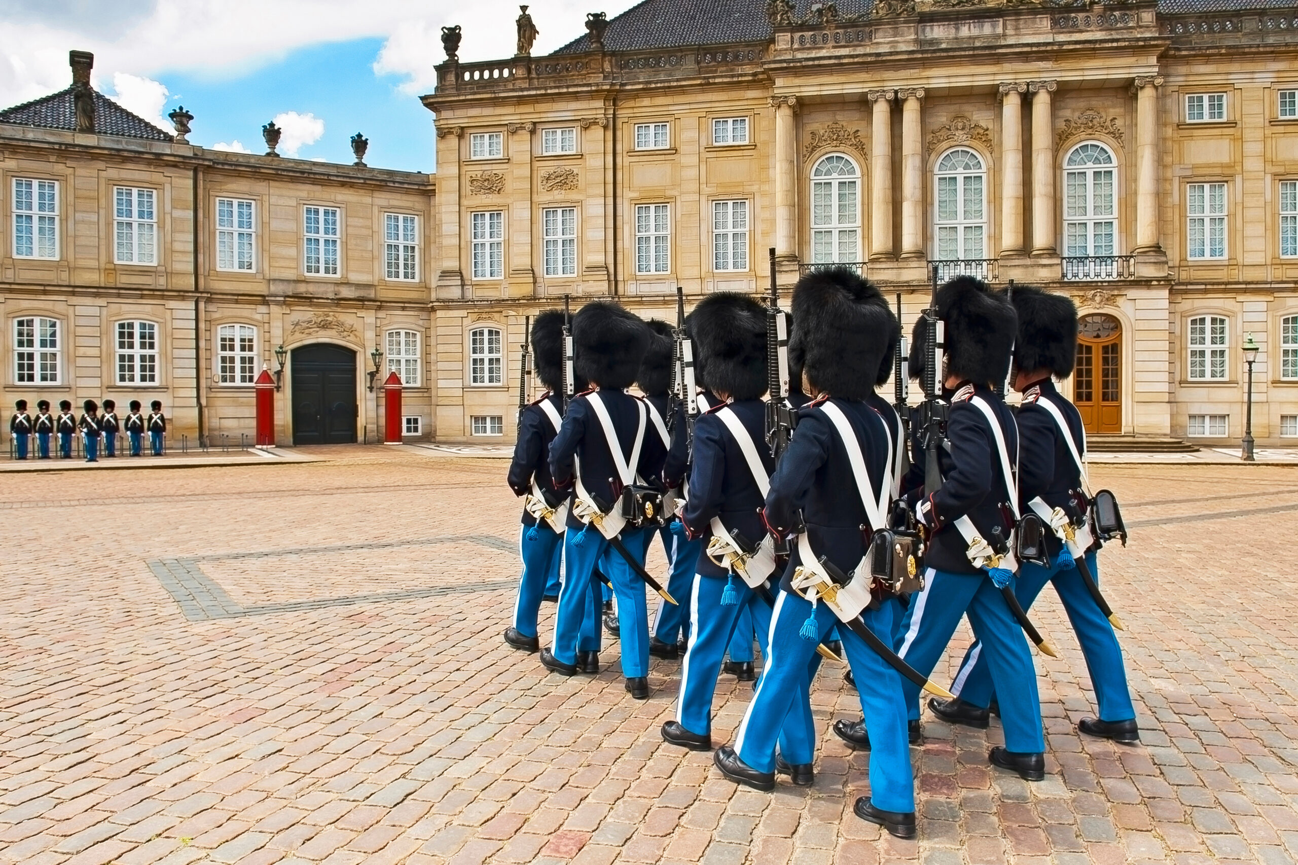 Row of ceremonial guards in blue uniforms marching with rifles in front of a beige neoclassical palace on a cobblestone square.