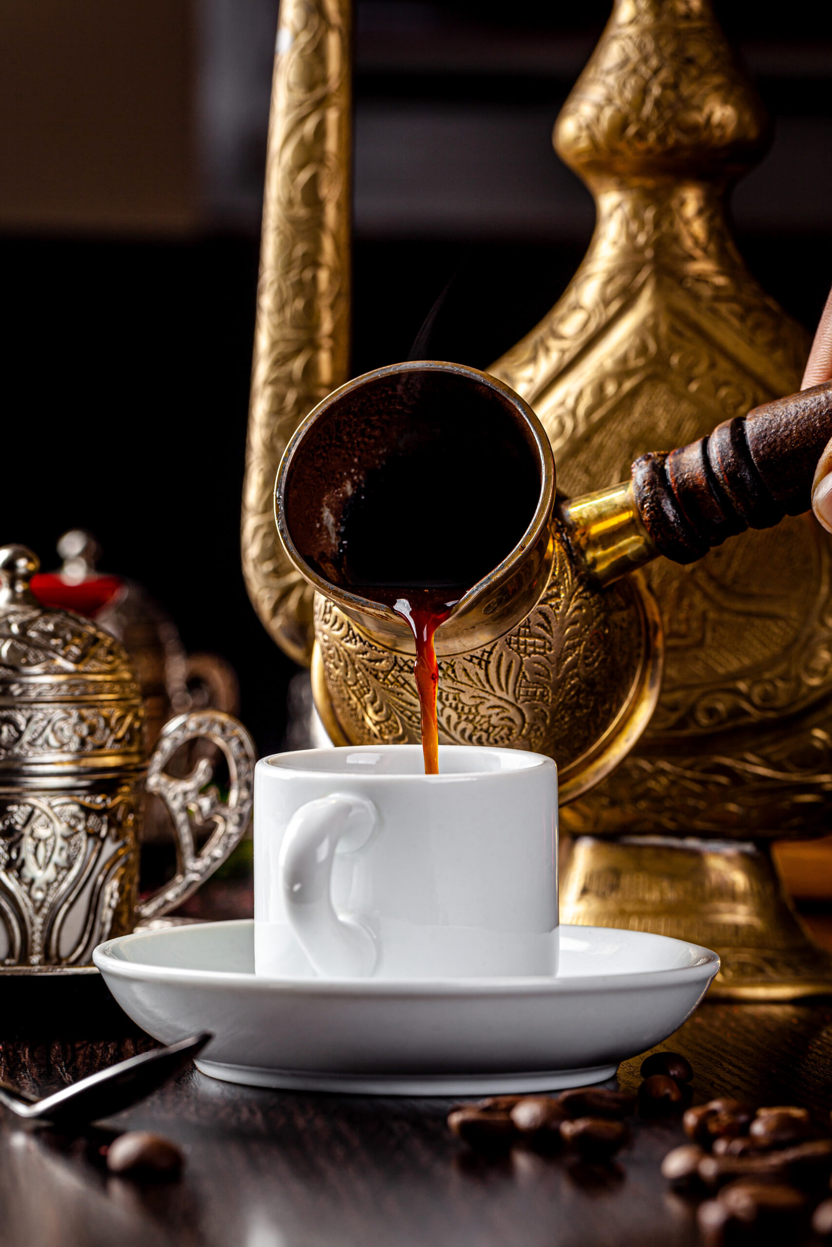 Close-up of dark coffee pouring from an ornate brass cezve into a white cup on a saucer, with a decorative gold teapot in the background