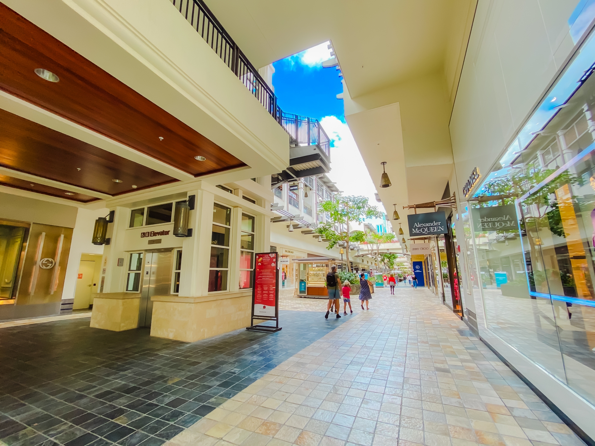 Open-air shopping street with storefronts, people walking, and a bright blue sky overhead.