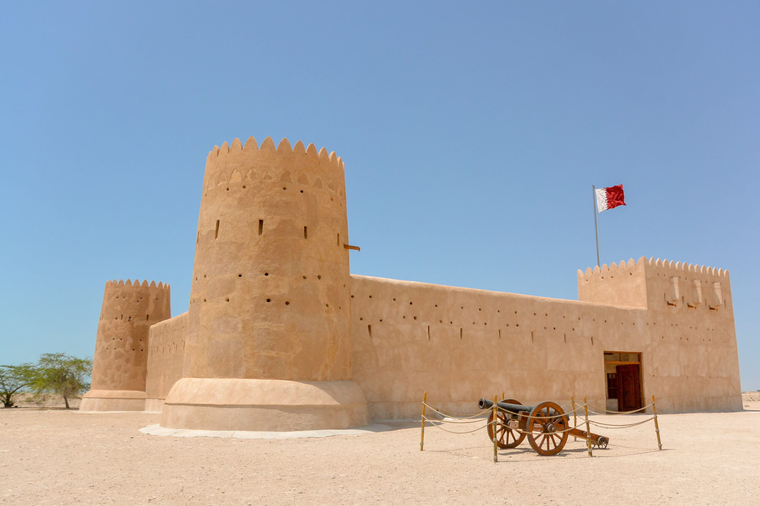 Desert fortress with a tall round tower and crenellated top, beige walls, a flag on a pole, and a cannon-wheel barrier in the foreground under a clear blue sky.
