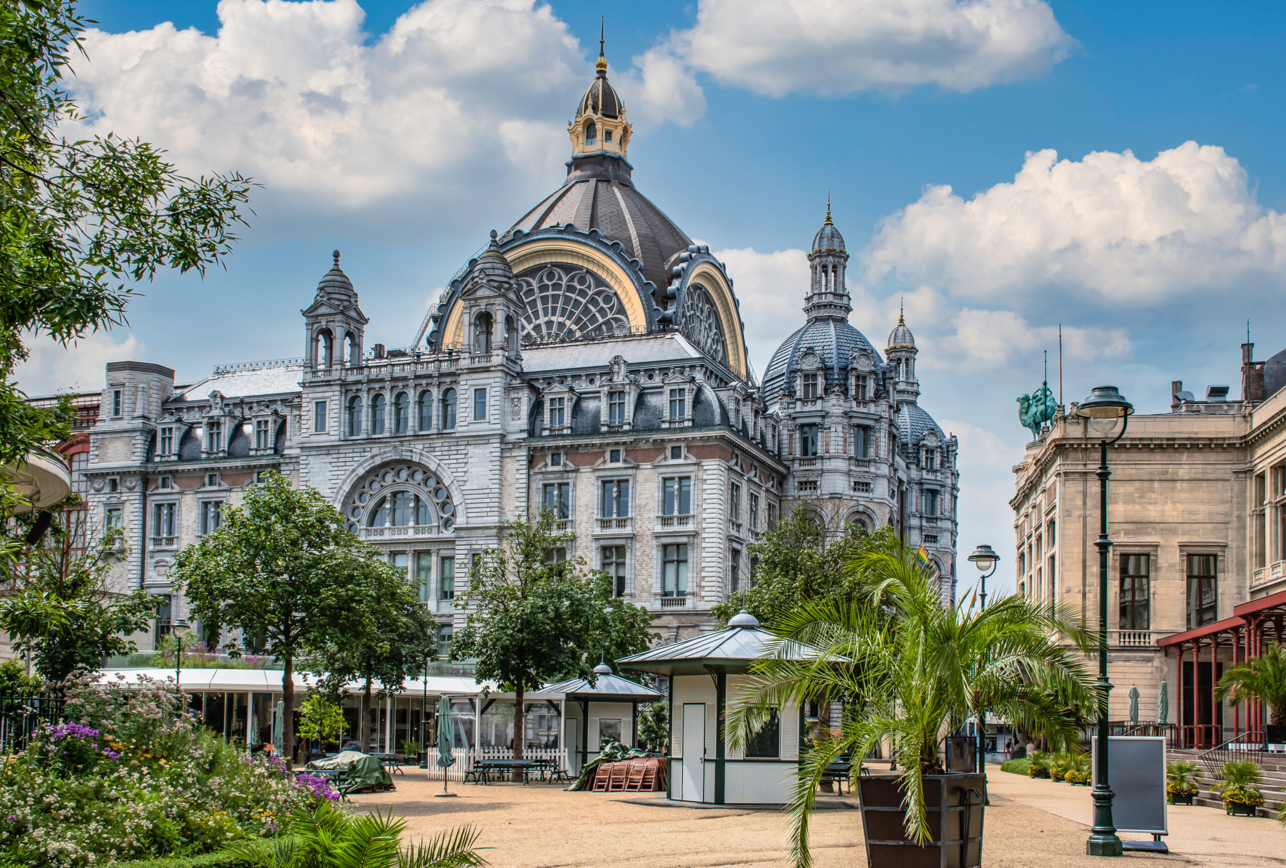 Ornate historic building with domed towers behind a sunny garden square and trees.