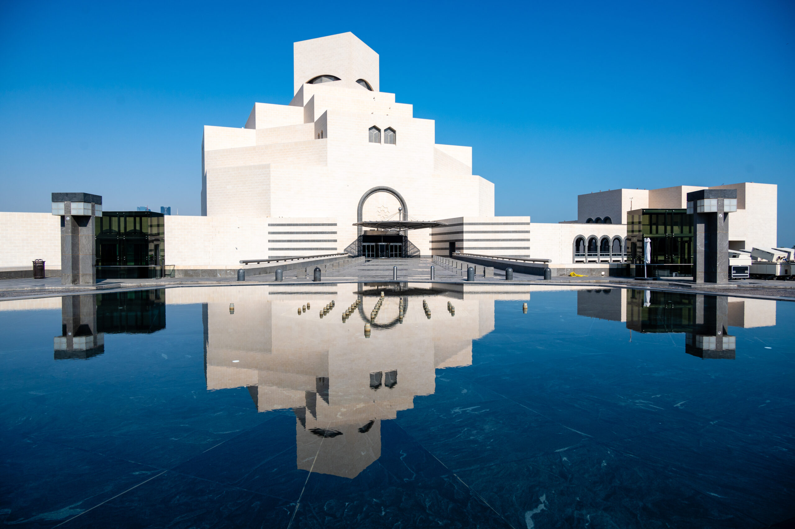 Modern white stone building with stepped tiers and arched windows, reflected in a shallow pool beneath a clear blue sky.