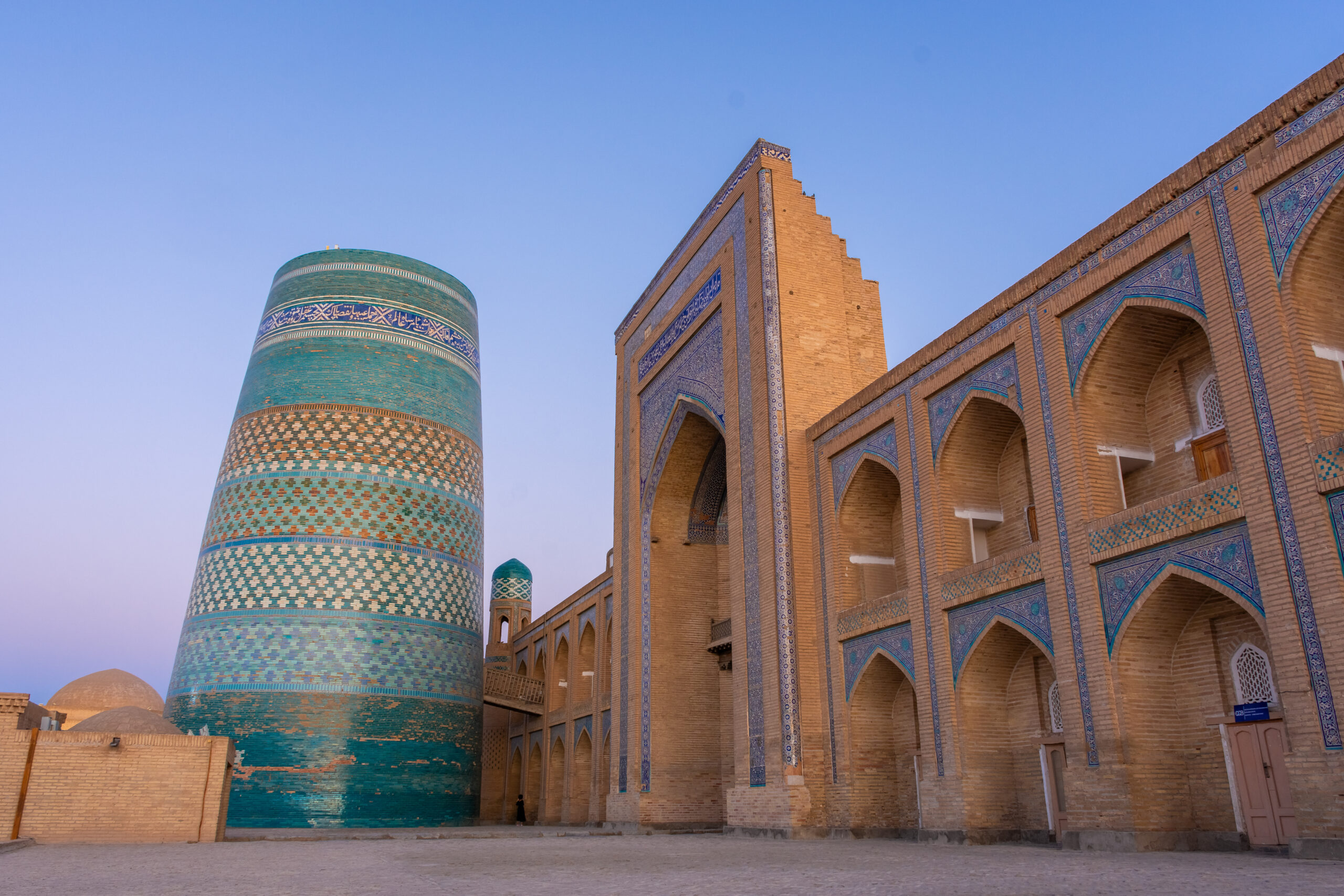 Turquoise tiled cylindrical minaret beside a brick building with blue mosaic arches under a clear blue sky.