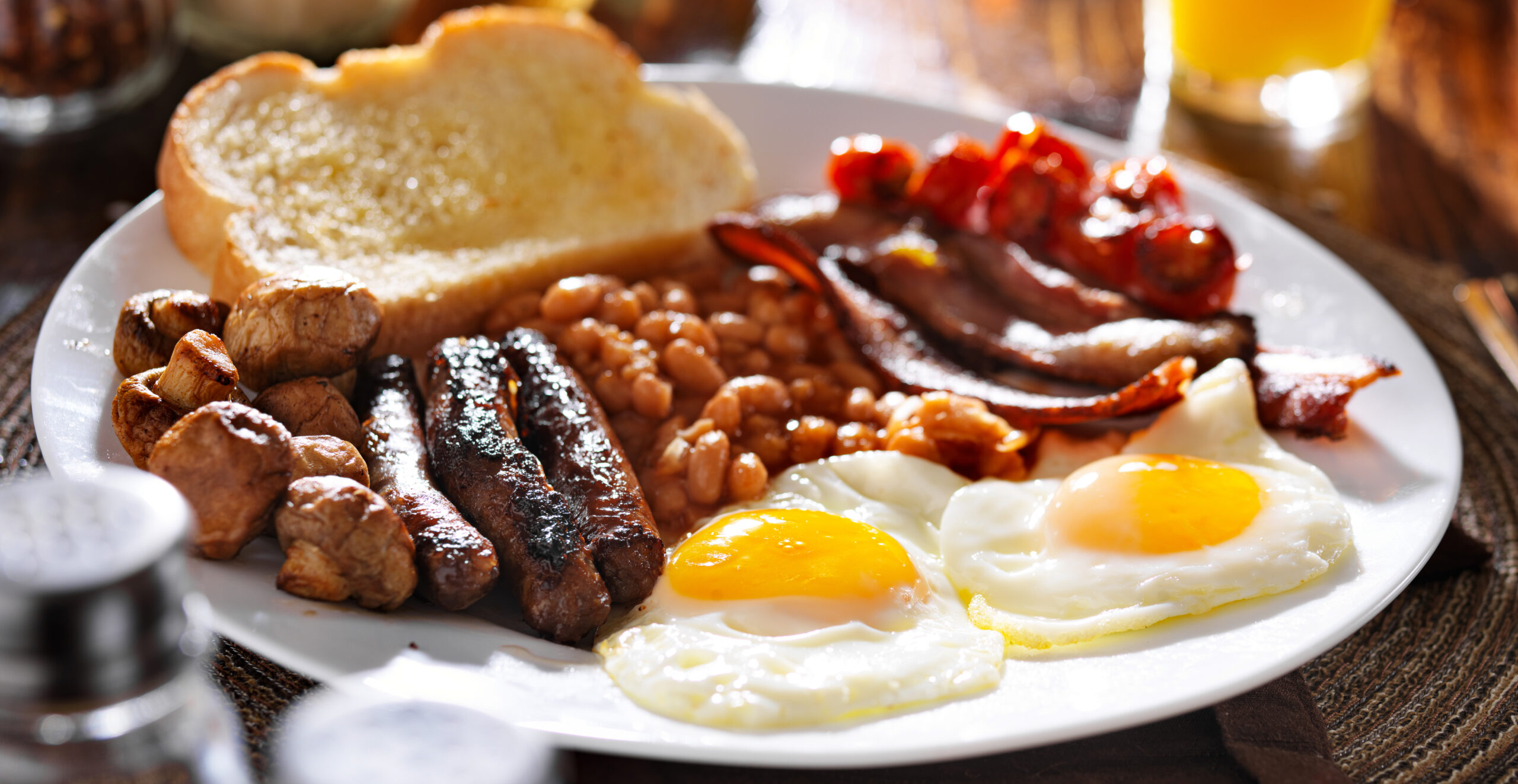 Plate of a full breakfast with sunny-side-up eggs, bacon, sausages, beans, mushrooms, tomatoes, toast, all on a white plate.