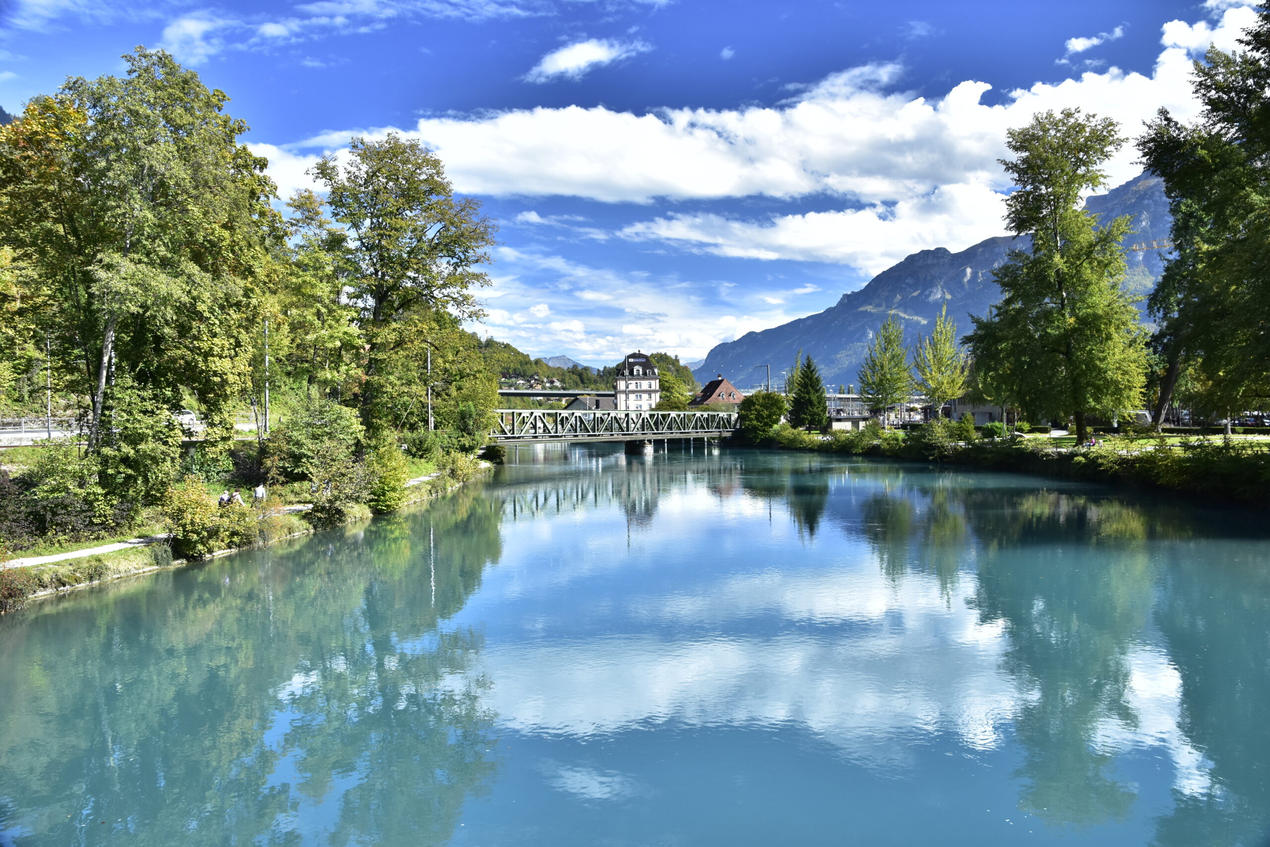Tranquil lakeside scene with green trees along the shore, a metal pedestrian bridge, and distant mountains under a blue sky with white clouds.
