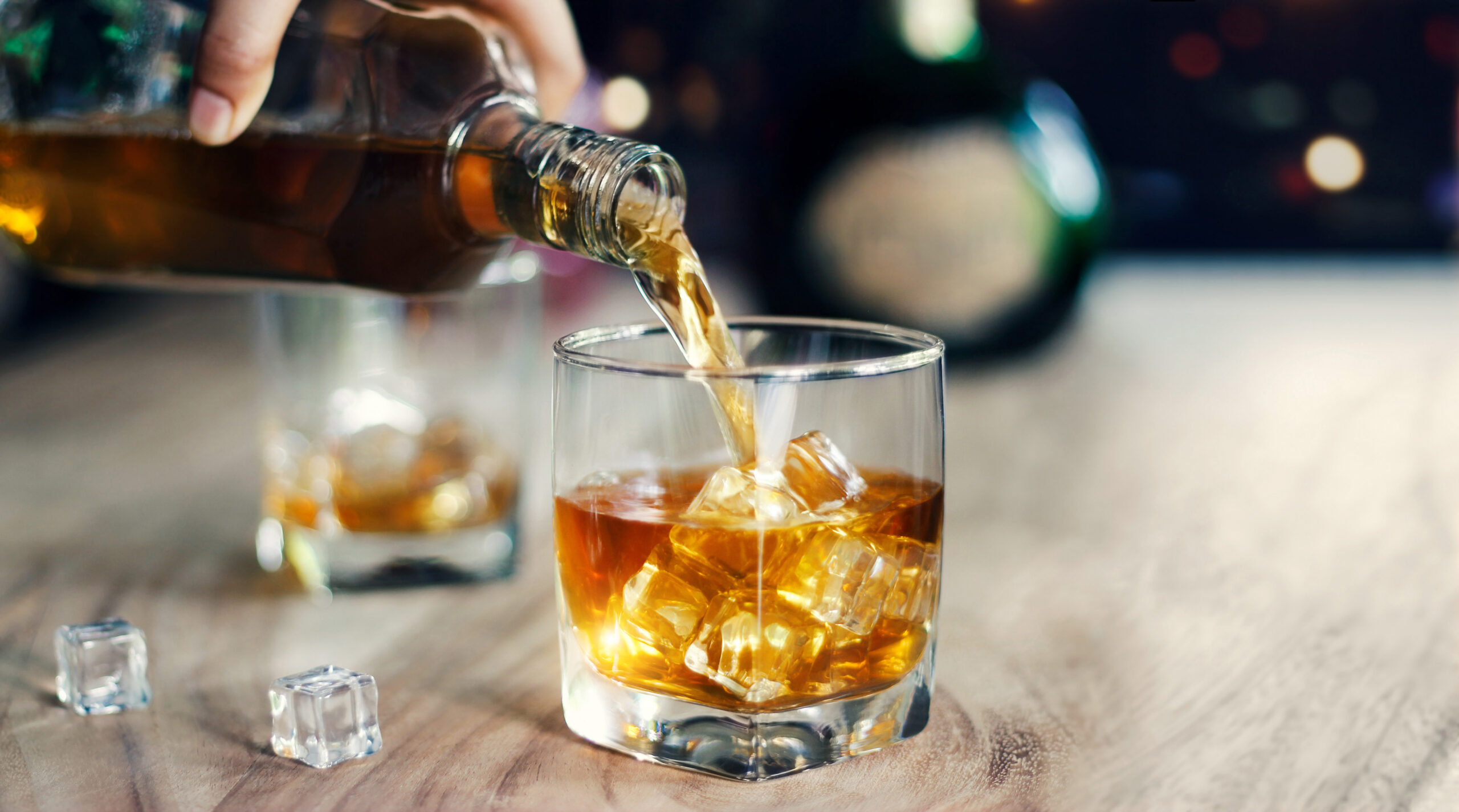 Whiskey being poured from a bottle into a glass with ice on a wooden table, with a blurred background.