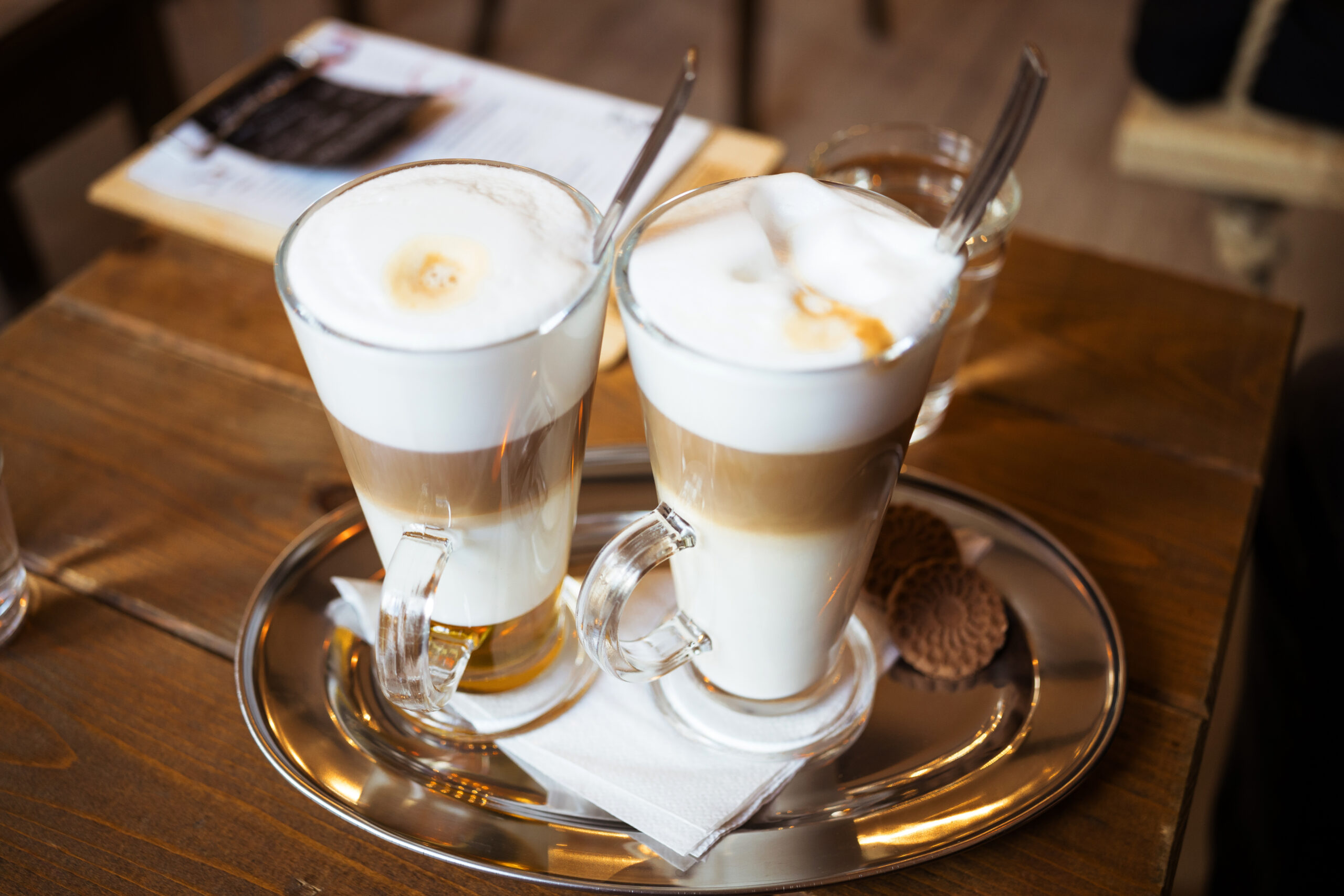 Two frothy coffee drinks in glass mugs on a silver tray, with spoons and a cookie on a wooden table.