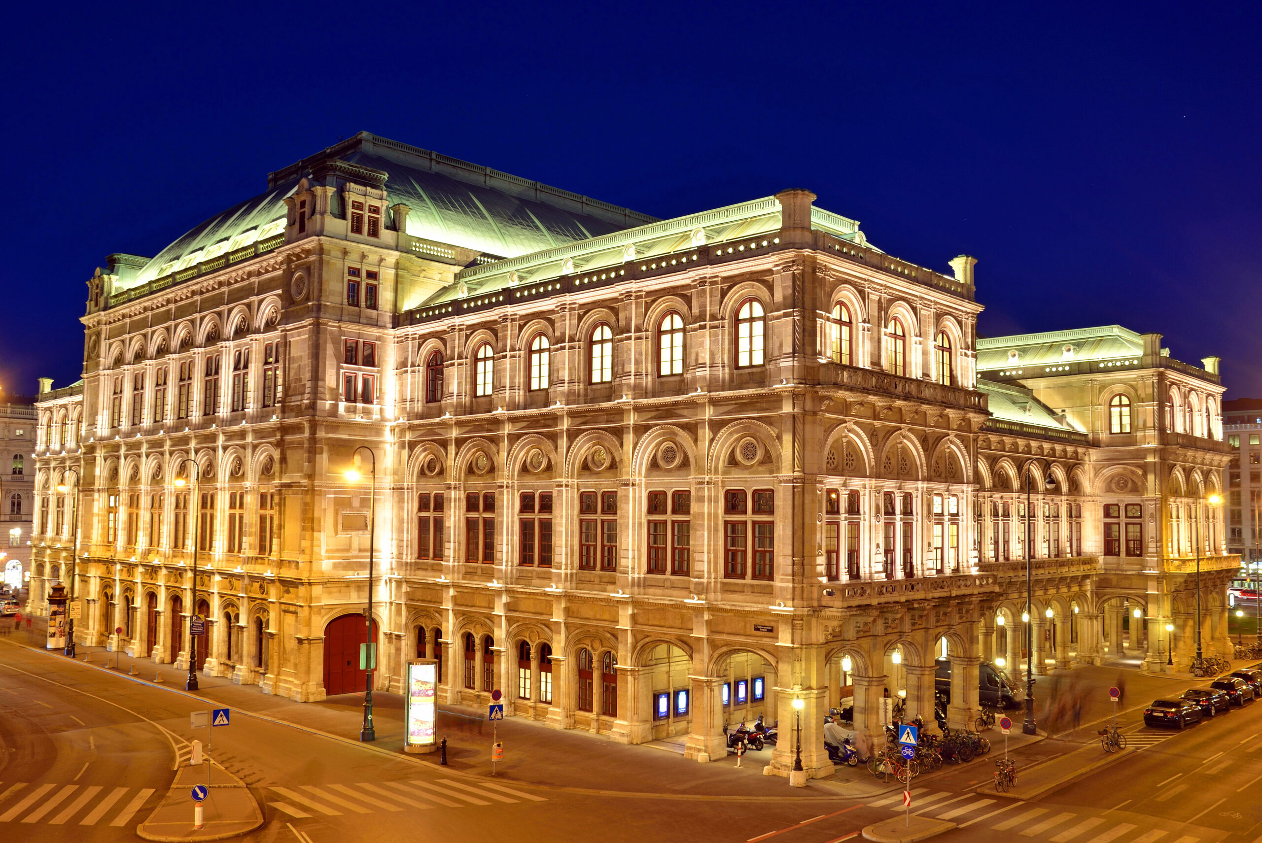 Night view of a historic illuminated building with arched windows and a domed roof along a quiet street at dusk/dawn.