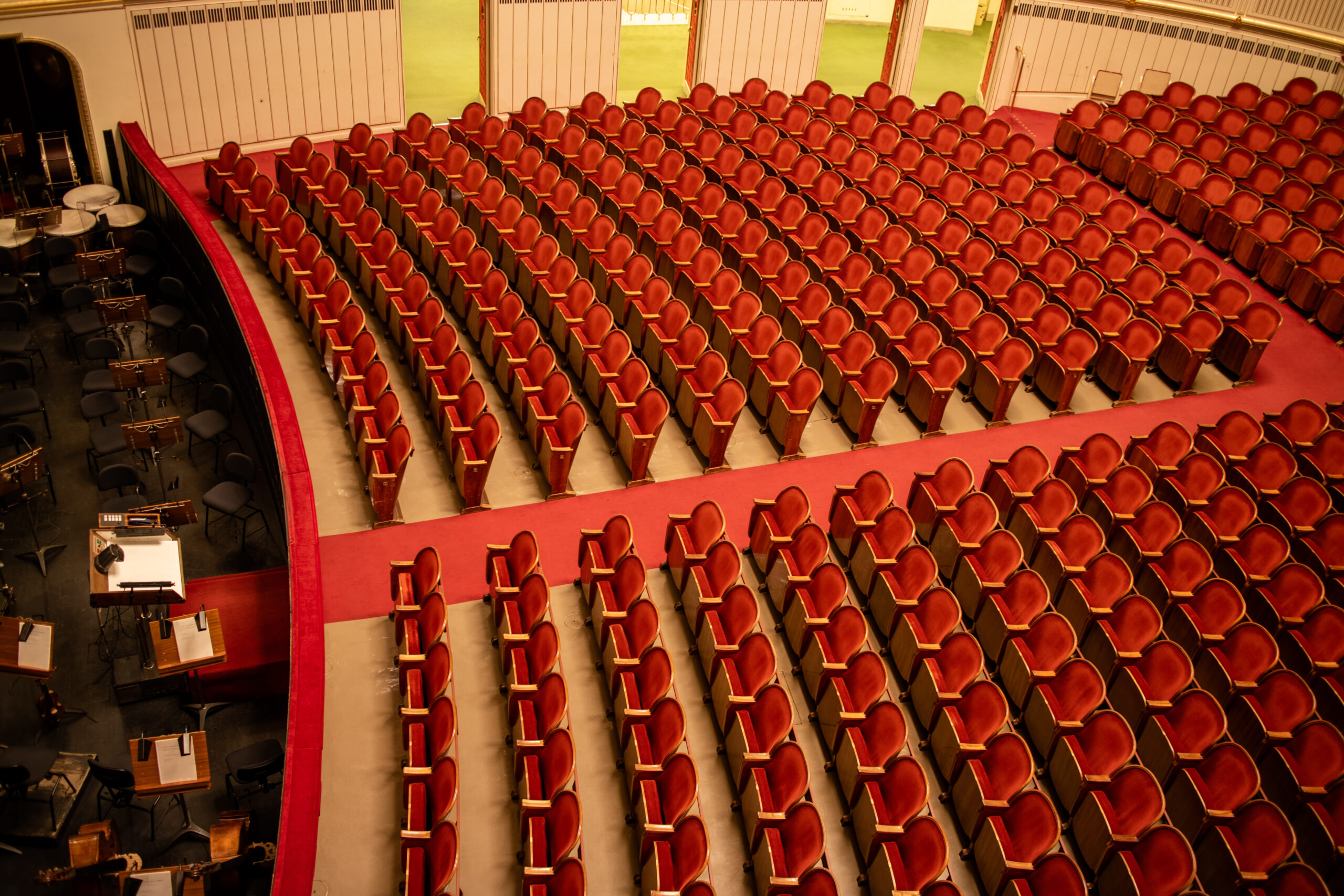 Top-down view of a large auditorium filled with red seats and a black orchestra pit to the left, instruments visible on stands.