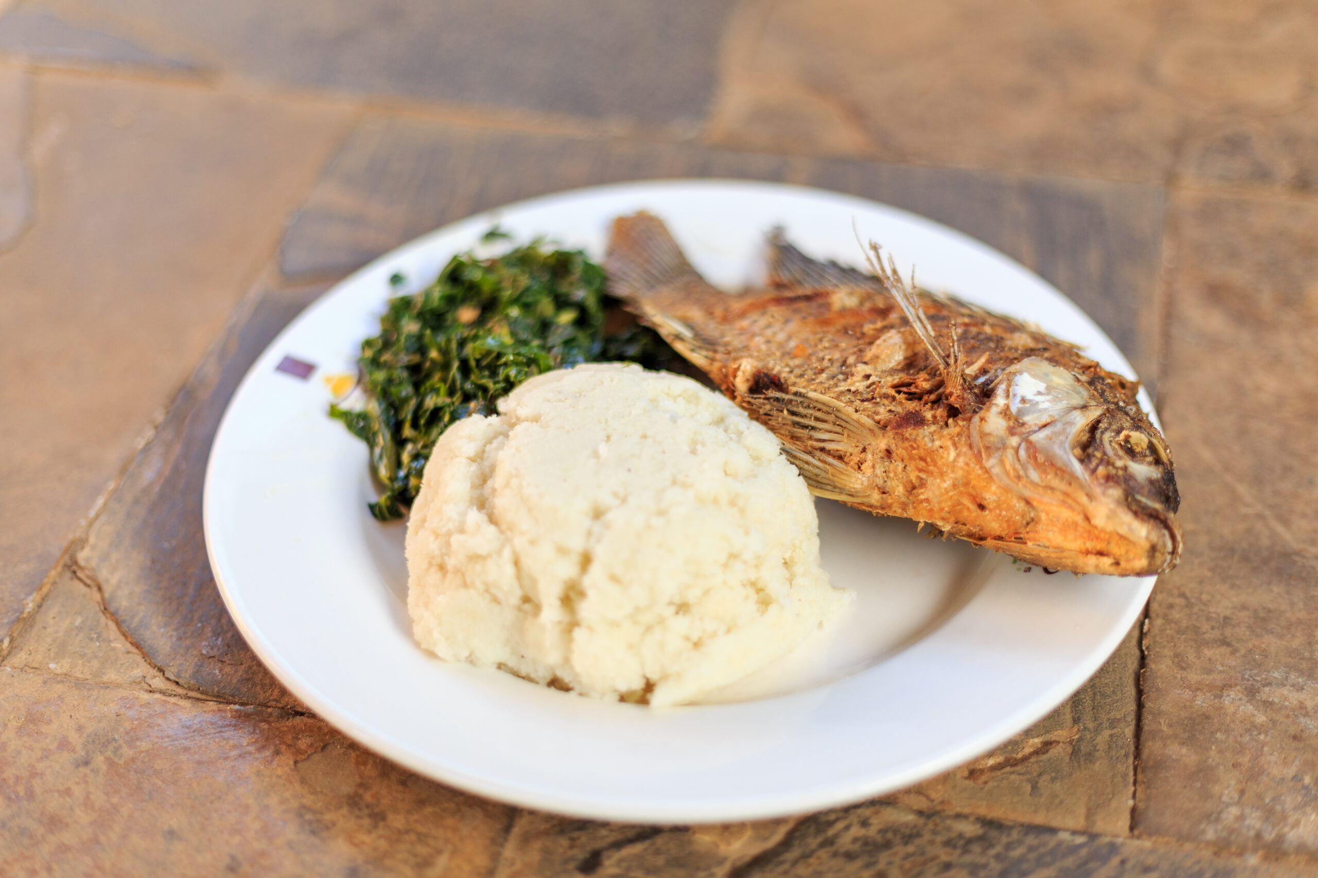Plate with fried fish, mashed potatoes, and sautéed greens on a rustic wooden table.