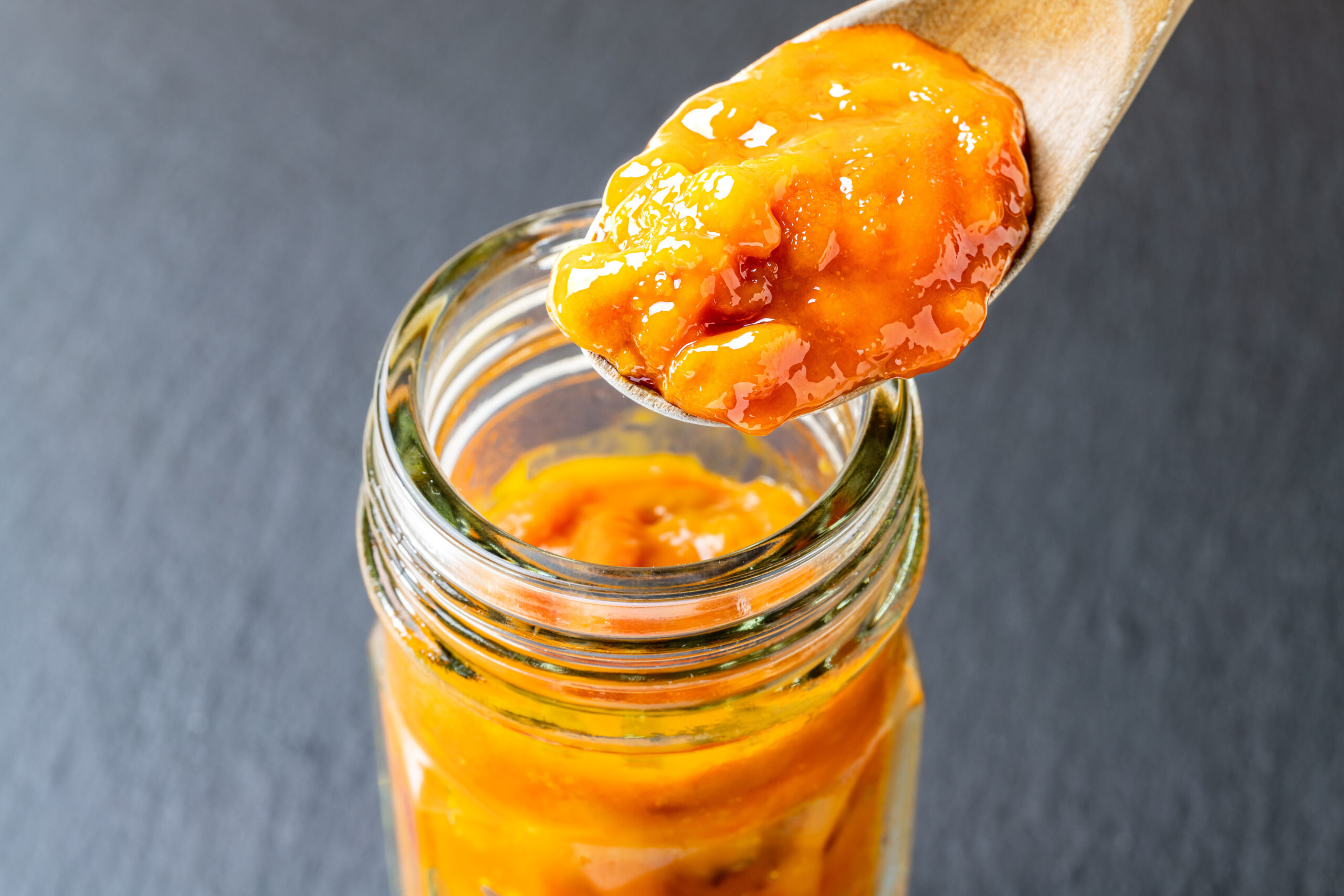 Close-up of orange jam on a wooden spoon above a glass jar, with jam dripping and glossy texture.