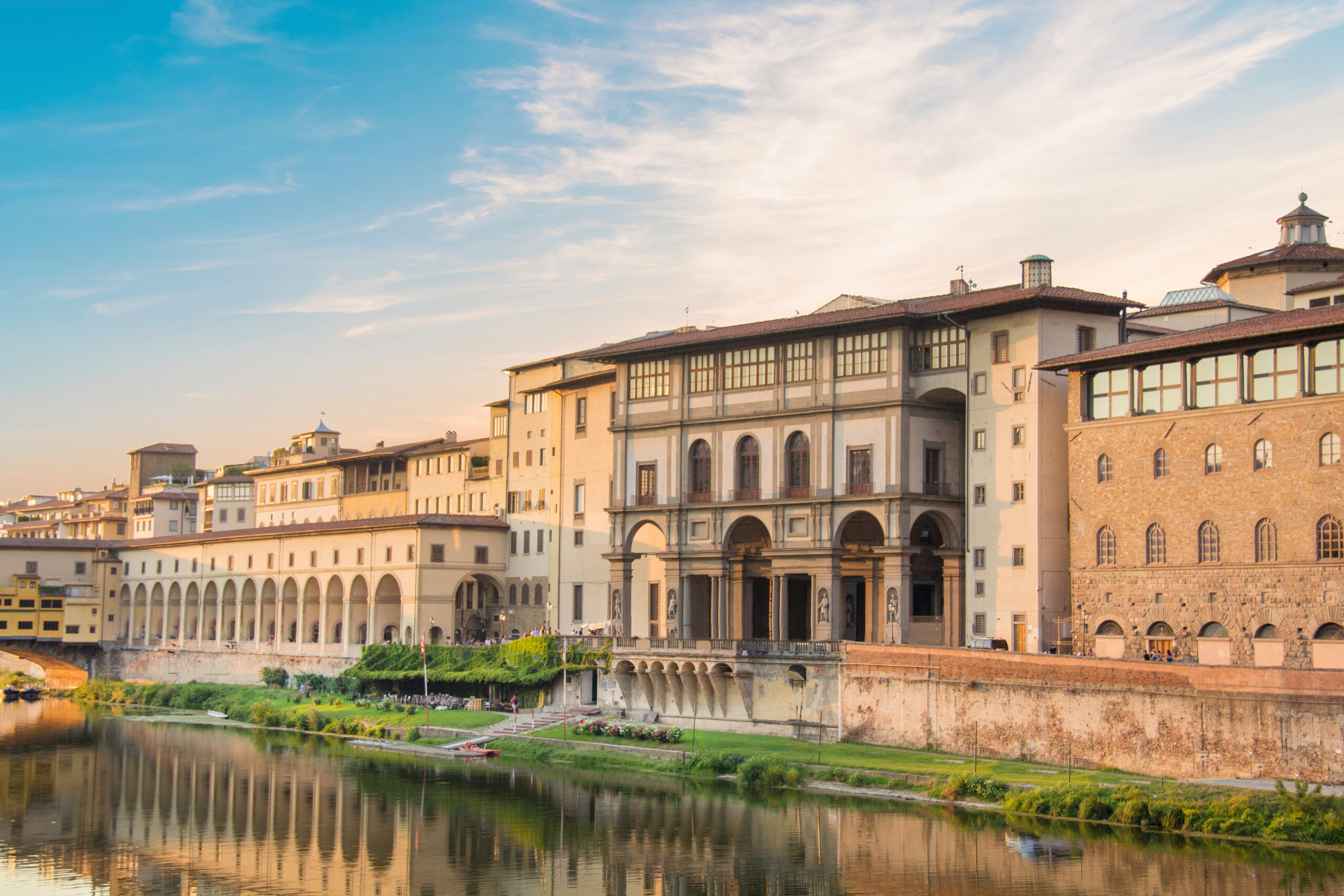 Historic riverside buildings with arches along a calm waterfront at sunset in warm light.