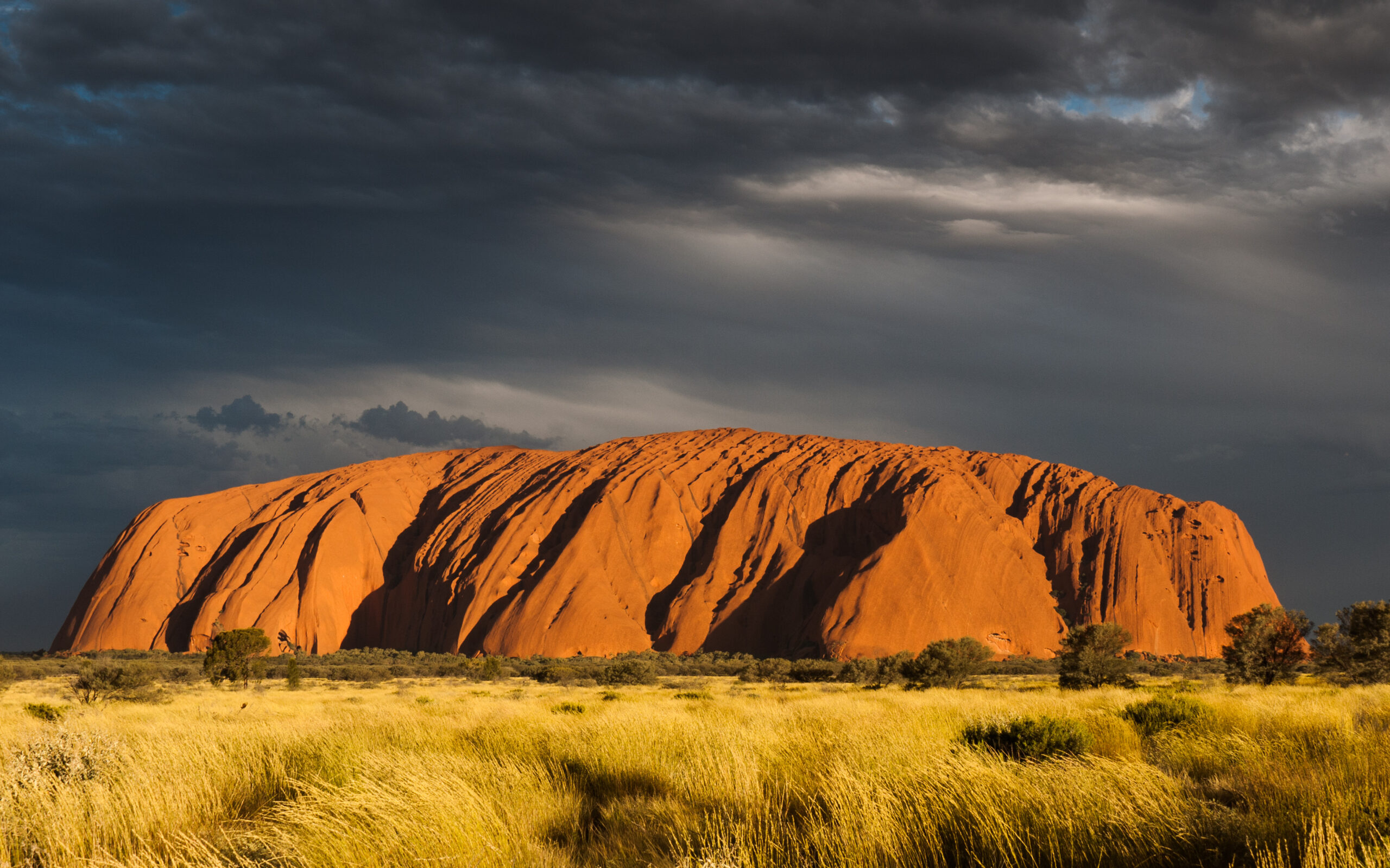 Sunlit orange rock formation rising from a flat desert plain under a dark, dramatic sky with scattered clouds.