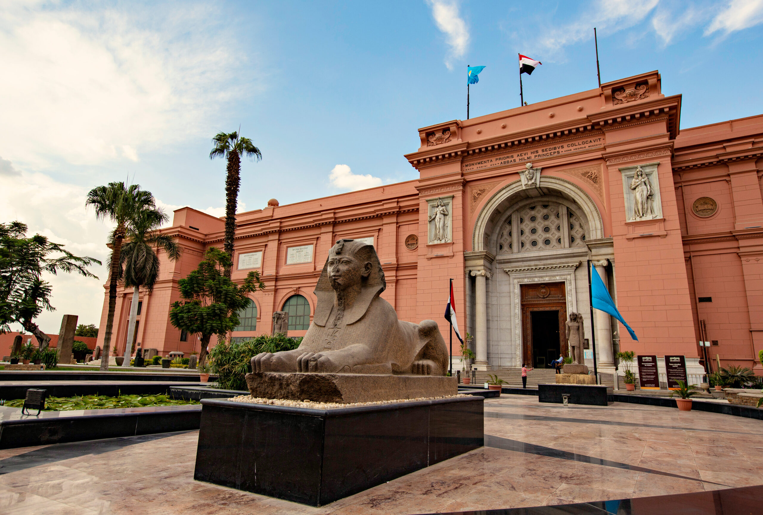 Pink neoclassical building with stone statues, flags, palm trees, and a sphinx sculpture in a plaza under a blue sky.