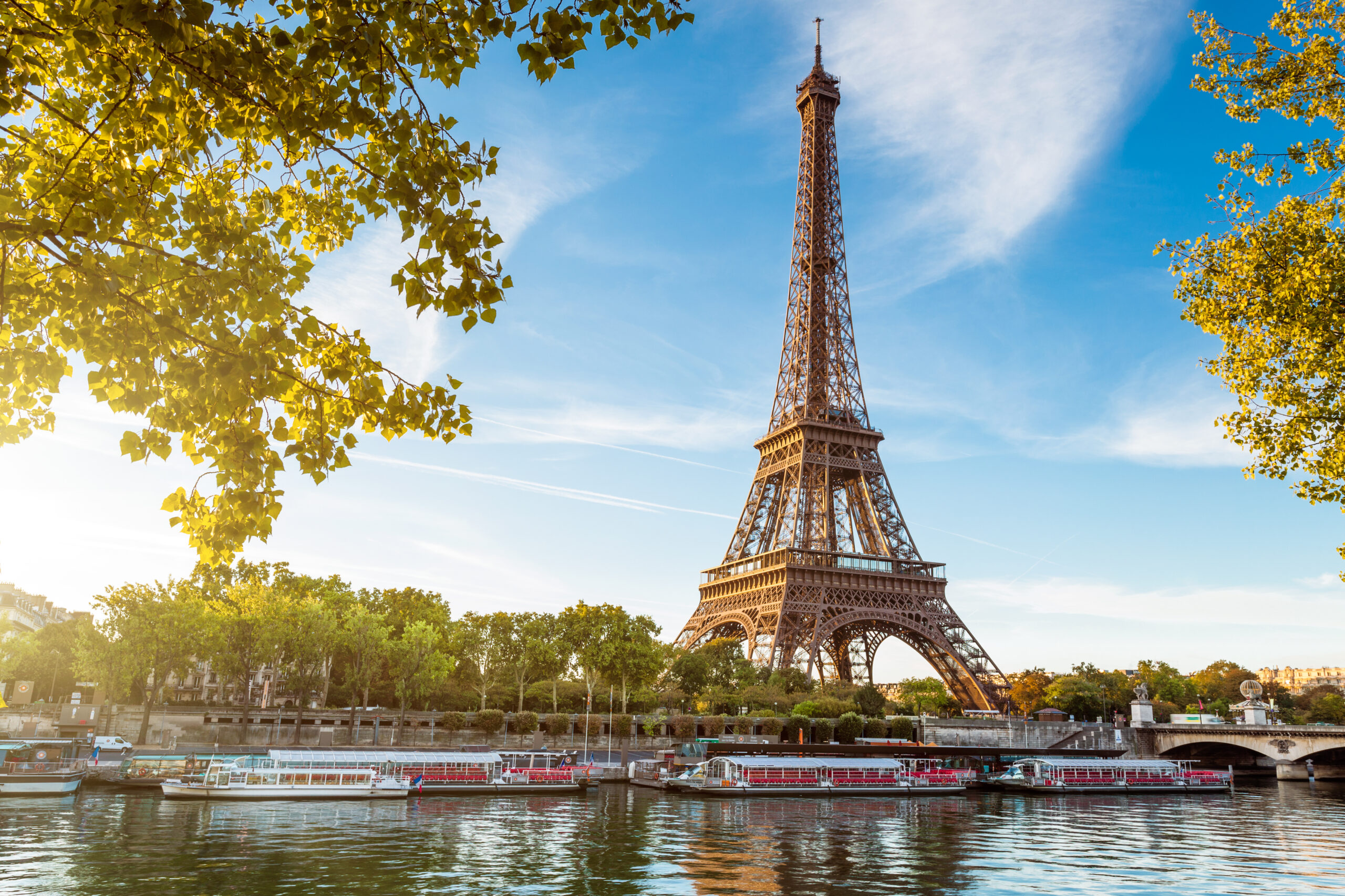 Eiffel Tower beside the Seine with boats on the river and green trees framing the scene under a clear blue sky.