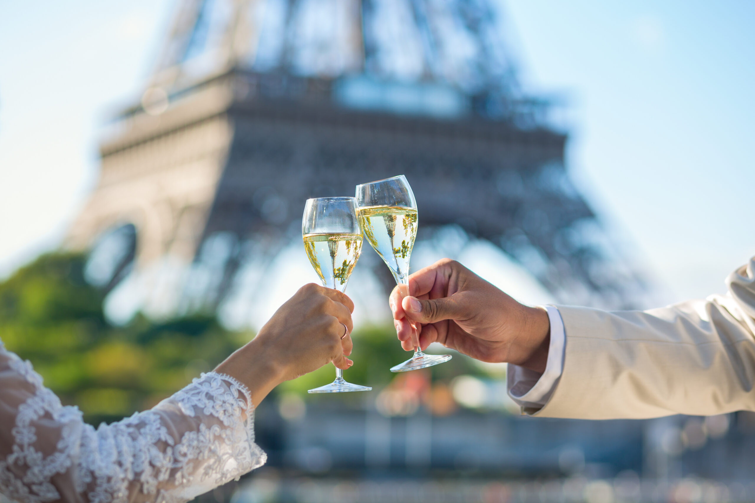 Two people clinking champagne flutes in a celebratory toast, with a blurred outdoor background and lace sleeves visible