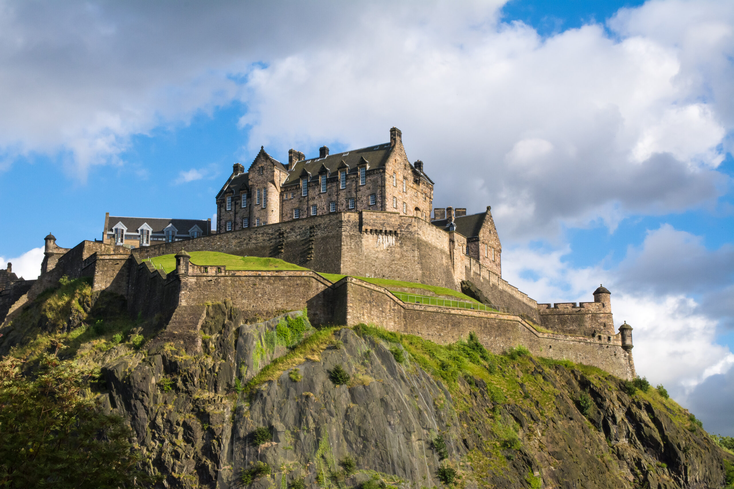 Stone castle atop a rocky cliff with green slopes and a blue sky behind it.