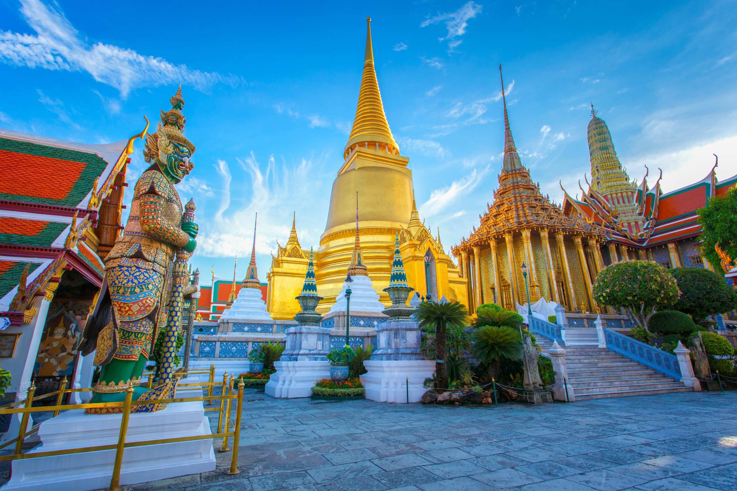 Golden central stupa with a tall spire at a Buddhist temple, surrounded by ornate gilded buildings and statues under a bright blue sky
