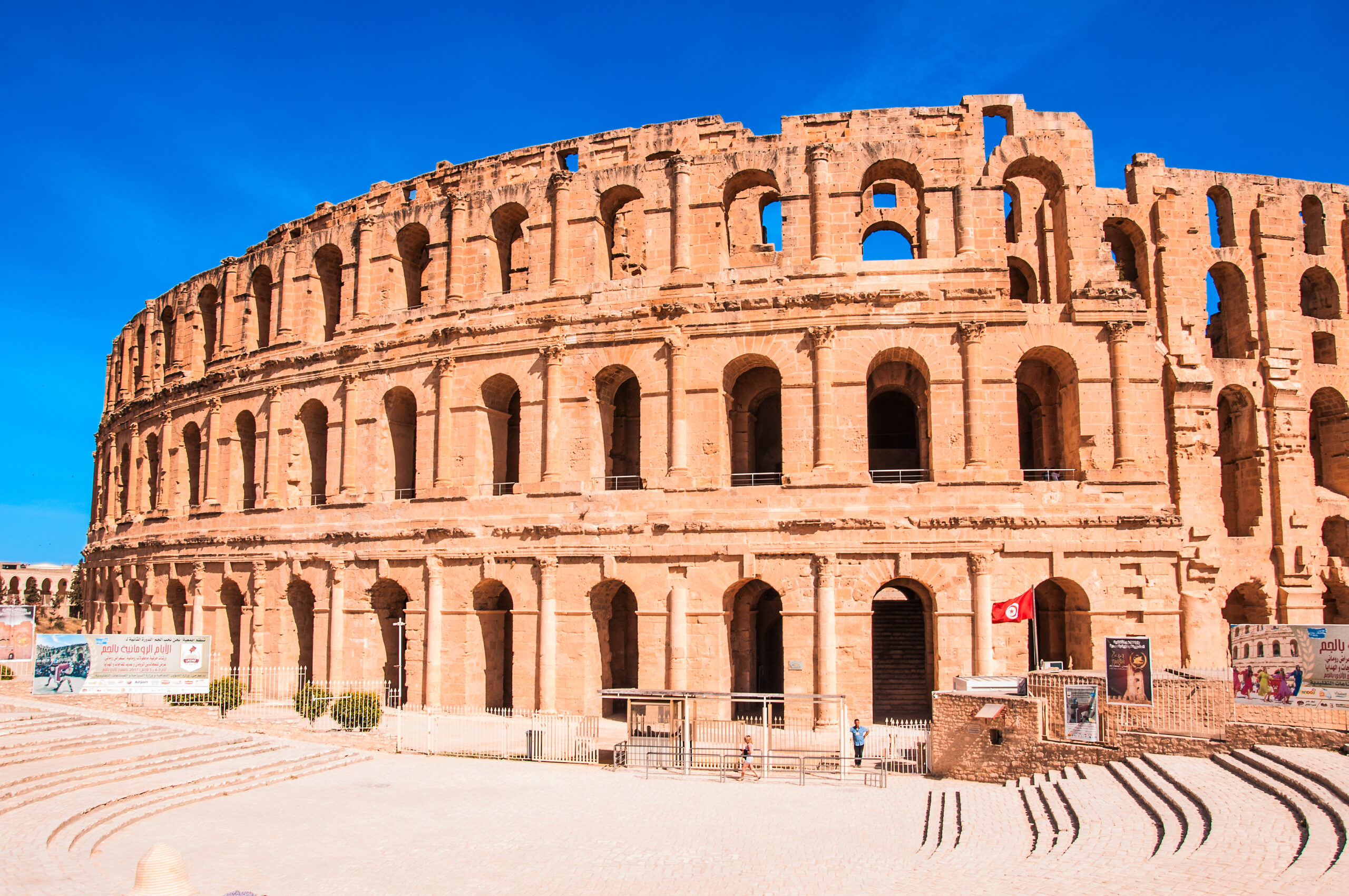 Exterior view of the ancient Roman Colosseum with arches against a bright blue sky.
