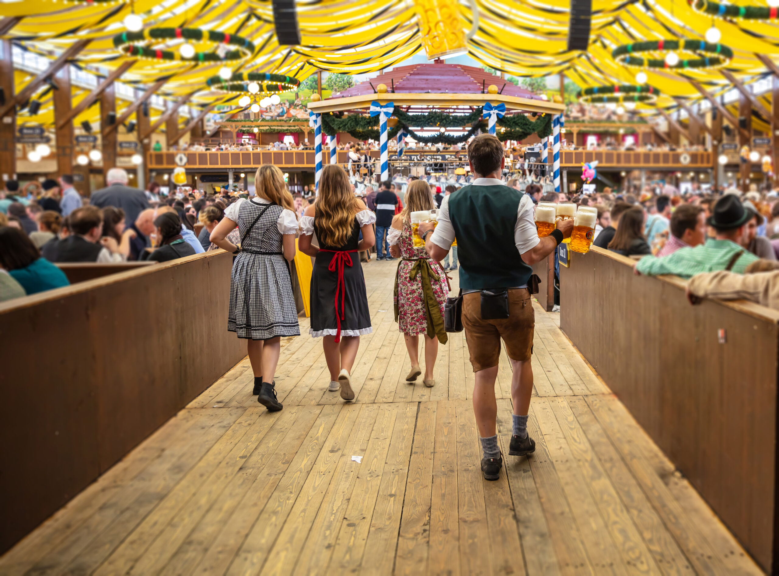 People in dirndl outfits walk down a wooden festival hall, lifting large beer steins amid yellow decorations overhead.