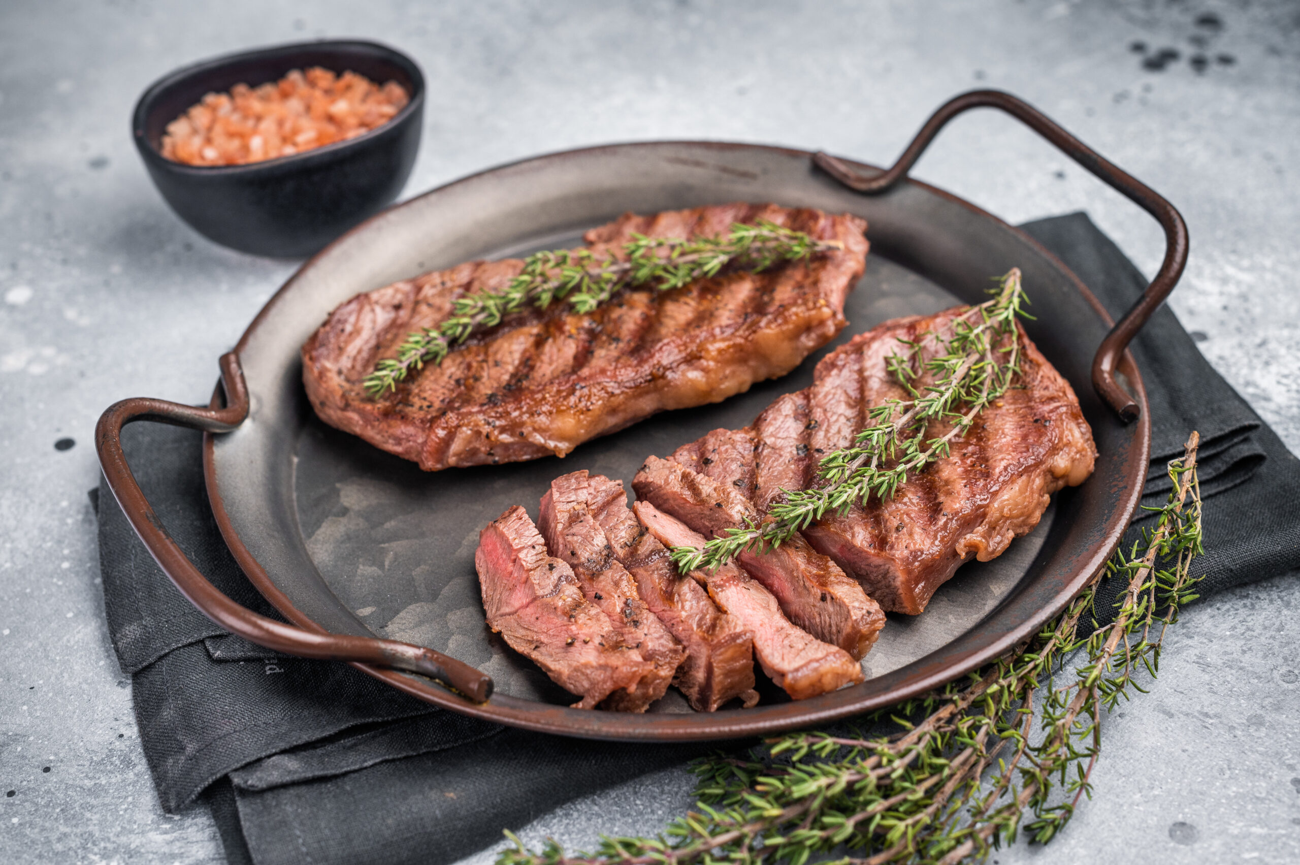 Slices of grilled steak on a cast-iron skillet, garnished with thyme, with a bowl of pink salt in the background.