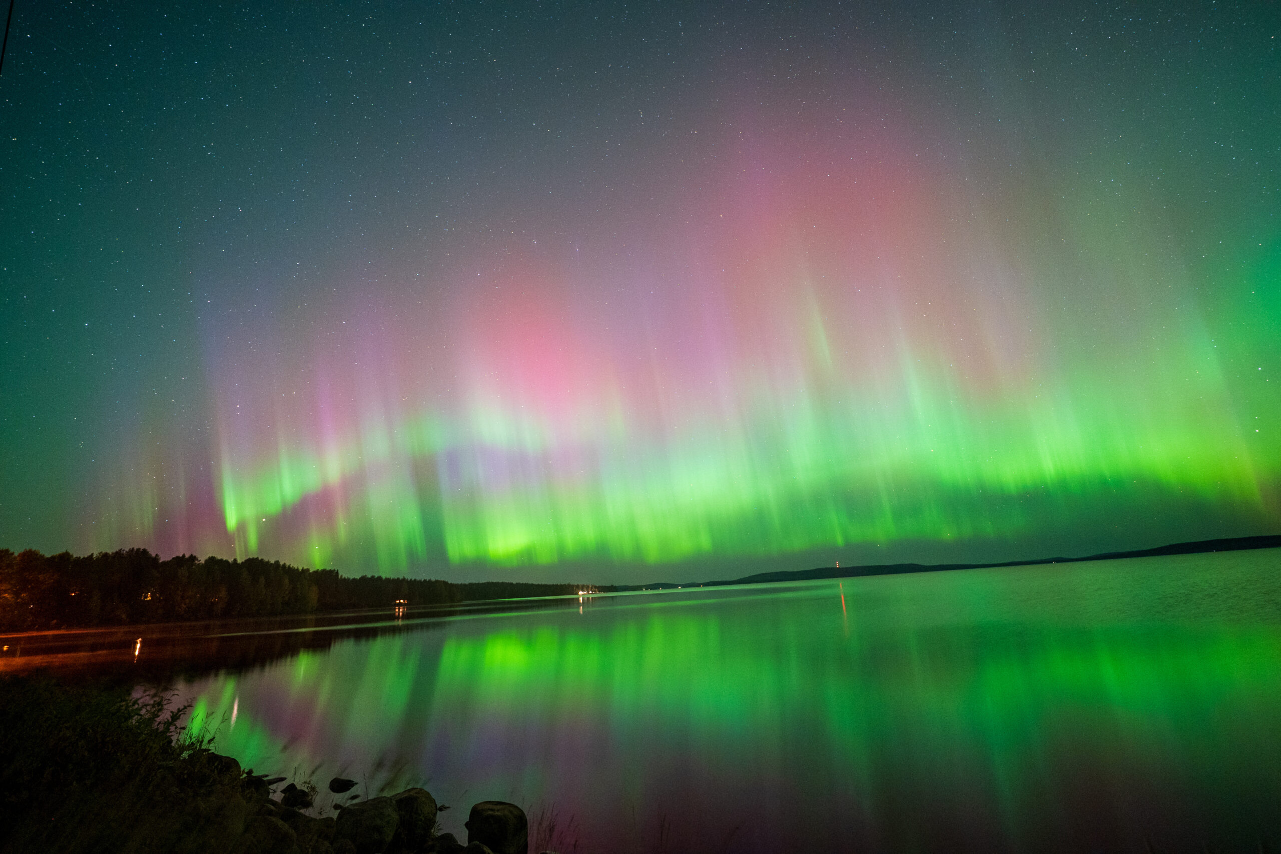 Aurora borealis lights the night sky over a calm lake, with green and pink hues reflecting on the water and dark shoreline at the bottom edge.