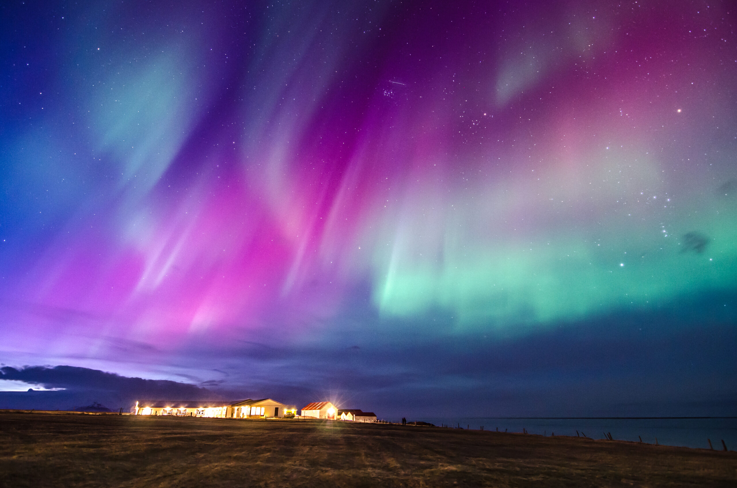 Aurora borealis coloring the night sky above a row of small, illuminated houses by the coast.